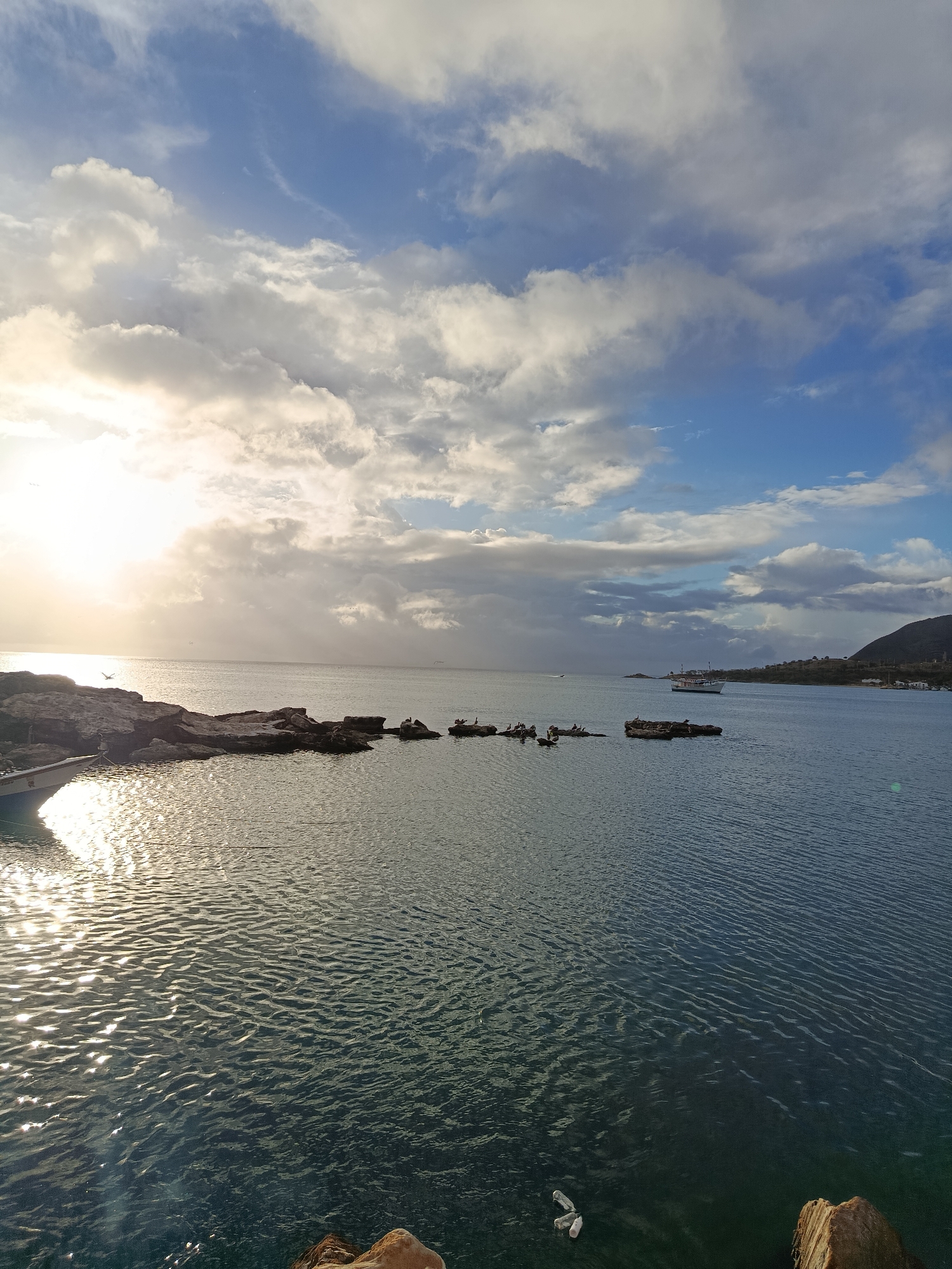 The image shows a body of water, likely a bay or harbor, under a partly cloudy sky. The sun is visible on the left side of the frame, casting a glare on the water's surface. Several rocky outcrops are present in the water, some with birds perched on them. A small boat is docked on the left, and a larger vessel is visible in the distance. A hilly coastline with some buildings can be seen on the right. The scene suggests a coastal environment, possibly a port or a scenic overlook in a city like Juan Griego, Venezuela, known for its historical importance and coastal views.