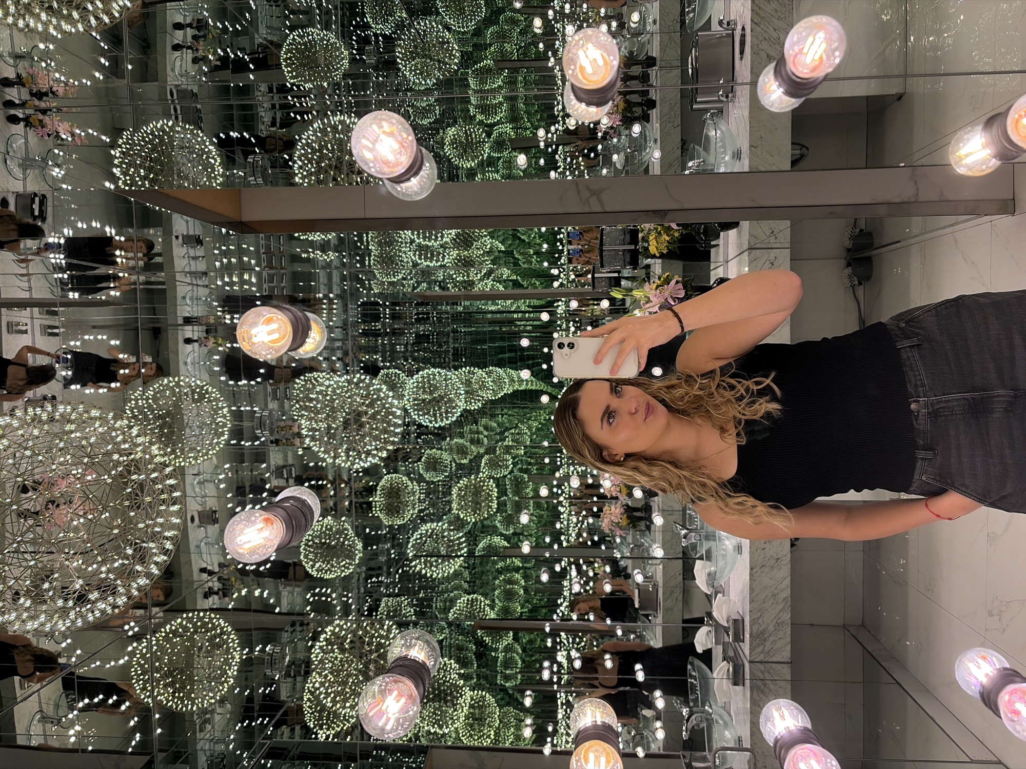 A fair-skinned woman with light brown hair is taking a photograph of herself in a room with mirrors. She is wearing a black shirt and black jeans. The mirrors create an infinity effect, with reflections of lights and decorative spheres. The room appears to be a restroom with sinks and toiletries visible in the reflections. The location is unknown, as the city and specific location are not identified.