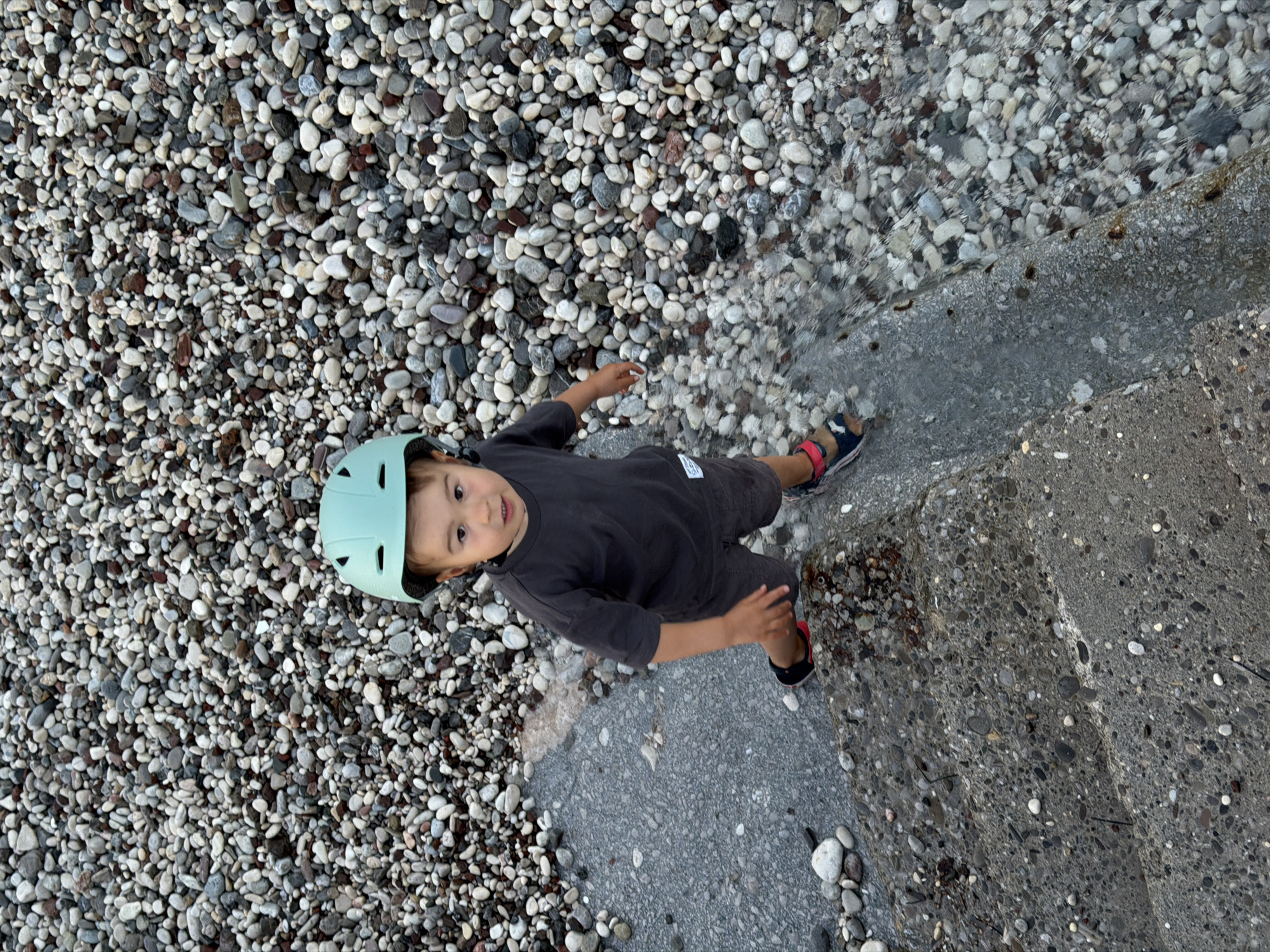 An overhead, slightly tilted shot captures a young child standing at the edge of a pebble beach, next to a section of concrete steps leading into shallow water. The child is wearing a light mint green helmet, a dark grey short-sleeved top with a small white tag on the left side, dark grey shorts, and blue and red sandals. They look directly upwards at the camera with a neutral, curious expression, their left arm slightly raised and bent at the elbow.
The ground is covered in a dense layer of small, smooth, multi-colored pebbles, ranging from white and light grey to darker grey and brown. On the right side of the frame, the pebbles meet clear, shallow water that partially covers the concrete steps. The child's right foot is placed on a concrete step, wet from the water, while their left foot is on the wet pebbles within the water. The water is calm and transparent, revealing the pebbles beneath its surface. The concrete structure appears to be a robust, grey wall or steps, also composed of aggregated stones, blending into the natural environment.
The scene depicts a sunny or brightly overcast daytime setting, likely at a beach or waterfront, consistent with the provided location context of Budva, Montenegro. The child appears to be engaged in gentle exploration or play by the water's edge. No discernible text is visible on any objects in the image.
