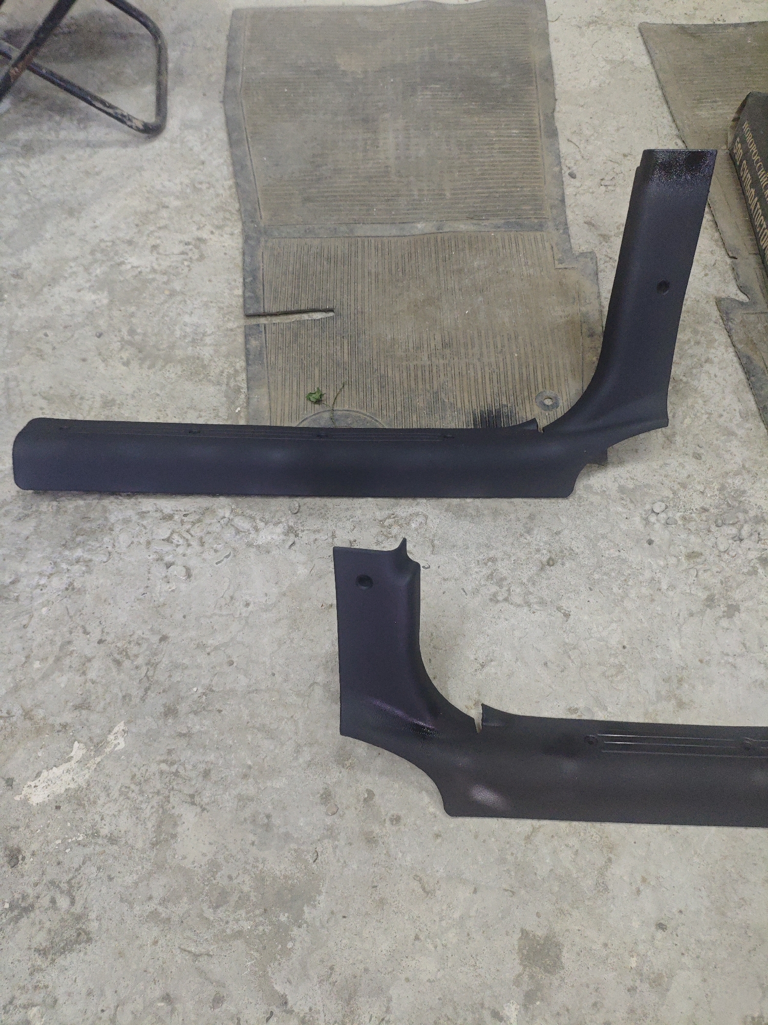 Two black plastic car interior trim pieces are laid out on a grey concrete floor. The pieces are curved and appear to be the A-pillar trim from a vehicle. One piece is longer than the other and is positioned horizontally in the upper portion of the image, while the shorter piece is positioned diagonally in the lower portion. A grey rubber mat is visible behind the longer trim piece. The concrete floor has some dirt and debris scattered on it. There is no indication of any specific location within Armavir, Russia, nor are there any people or actions depicted in the image.