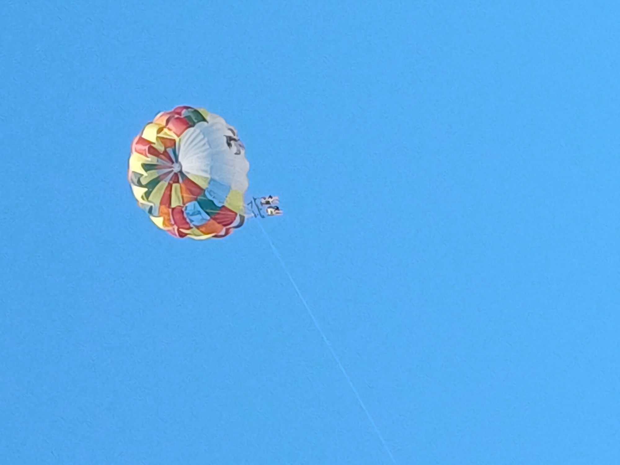 The image depicts a vibrant, multicolored parachute filled with air, soaring against a clear, bright blue sky. Attached to the parachute are two individuals suspended in a harness, appearing to be enjoying a parasailing adventure. A thin, taut rope extends diagonally from the parachute downwards and to the left, presumably connecting it to a boat or vehicle out of frame, which is pulling the parachute. The background is uniformly blue, indicating an open sky, likely over an oceanic or large body of water setting. The parachute exhibits a pattern of colored segments, including yellow, red, green, orange, and white. The overall scene captures a moment of leisure and outdoor recreation under fair weather conditions. There is no visible indication of the city of Sochi, Russia in this image.