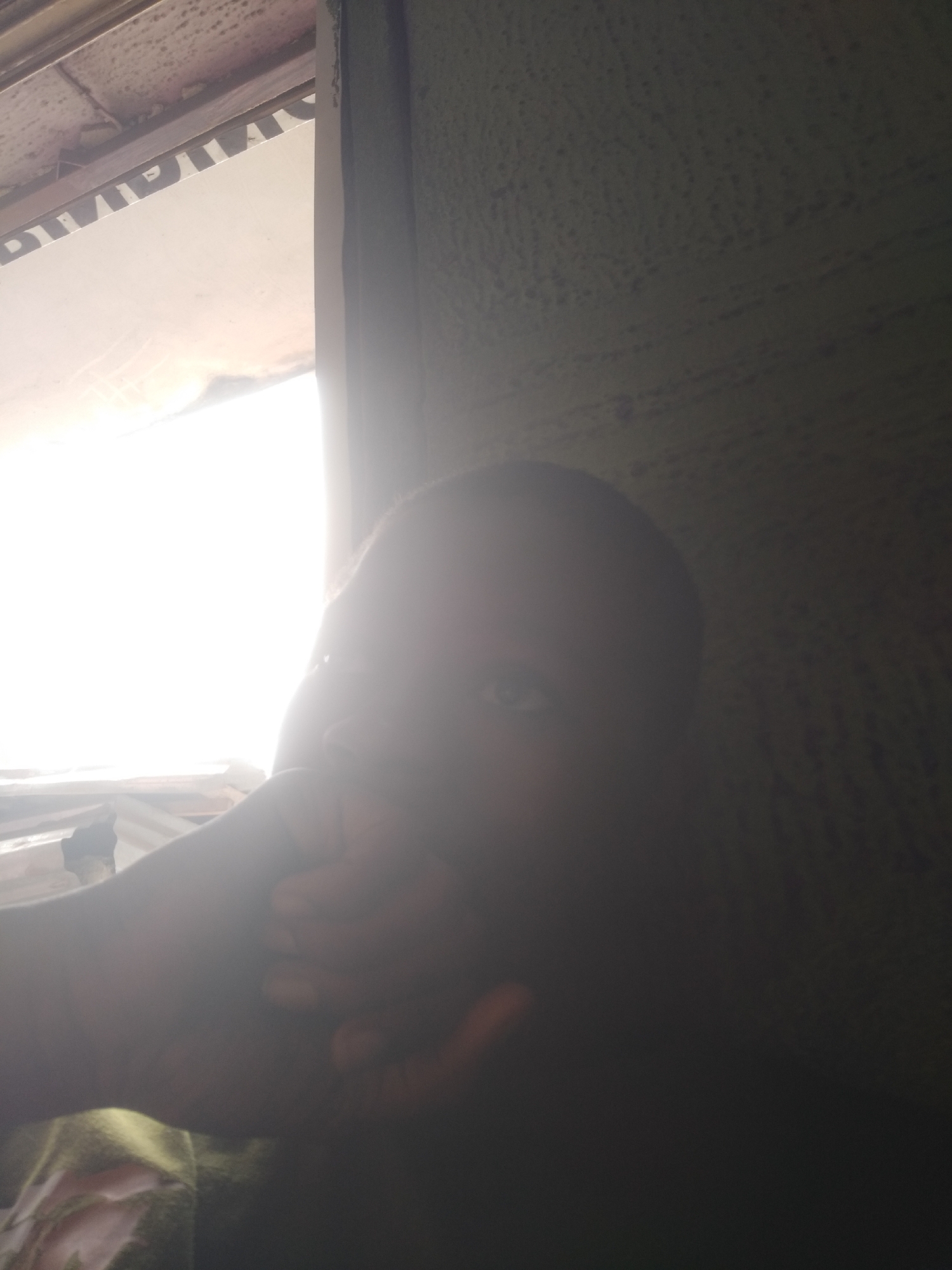 The image shows a person indoors, likely in a room in Shangisha, Nigeria. The person's face is partially obscured by their hand and is heavily shadowed, with a bright light source coming from a window to the left. The background wall appears textured. The subject is wearing a green shirt.