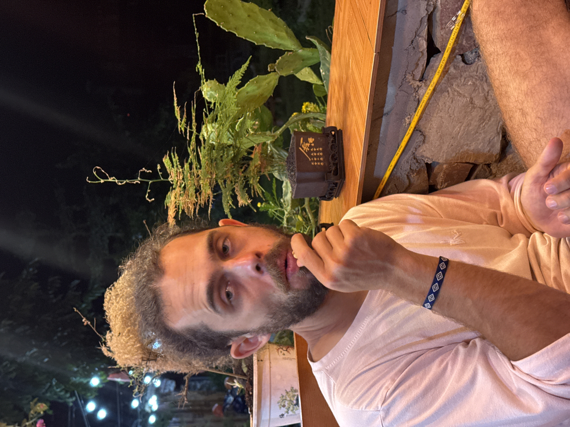 A man with curly hair and a beard is looking at the camera with his hand on his chin. He is wearing a light pink t-shirt and a blue bracelet with white diamond patterns. Behind him is a wooden structure with stone on the side, a cactus plant, and other greenery. In the dark background, there are blurred lights. The man is presumably in Mèo Vạc Commune, Vietnam.