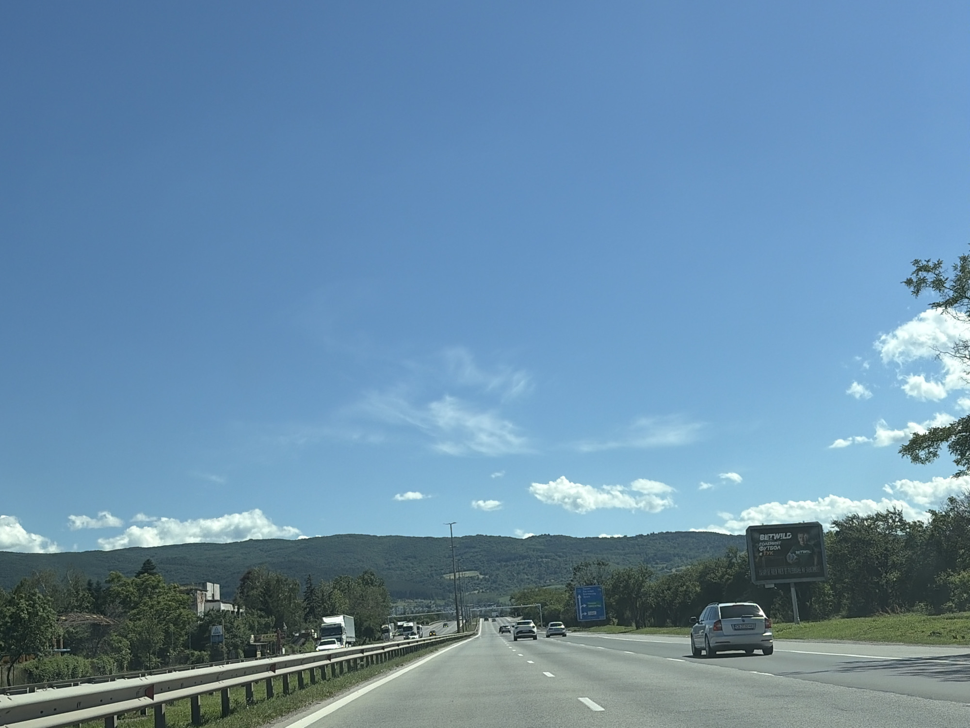 A bright, sunny day is depicted with a clear blue sky and scattered white clouds. The image appears to be taken from a vehicle traveling on a multi-lane highway. On the left side of the highway, there is a guardrail and a grassy embankment. Beyond that, there are trees and some buildings, including what looks like an industrial or abandoned structure. In the distance, a large, tree-covered mountain range stretches across the horizon.
On the right side of the highway, there is a grassy shoulder with more trees and vegetation. A billboard is visible in the distance, featuring an advertisement for "BETWILD" with text that appears to be in a foreign language. Several cars and trucks are on the highway, traveling in the same direction as the viewer. The road surface is asphalt, with clear lane markings. The overall impression is a rural or suburban highway environment under a fair weather sky.