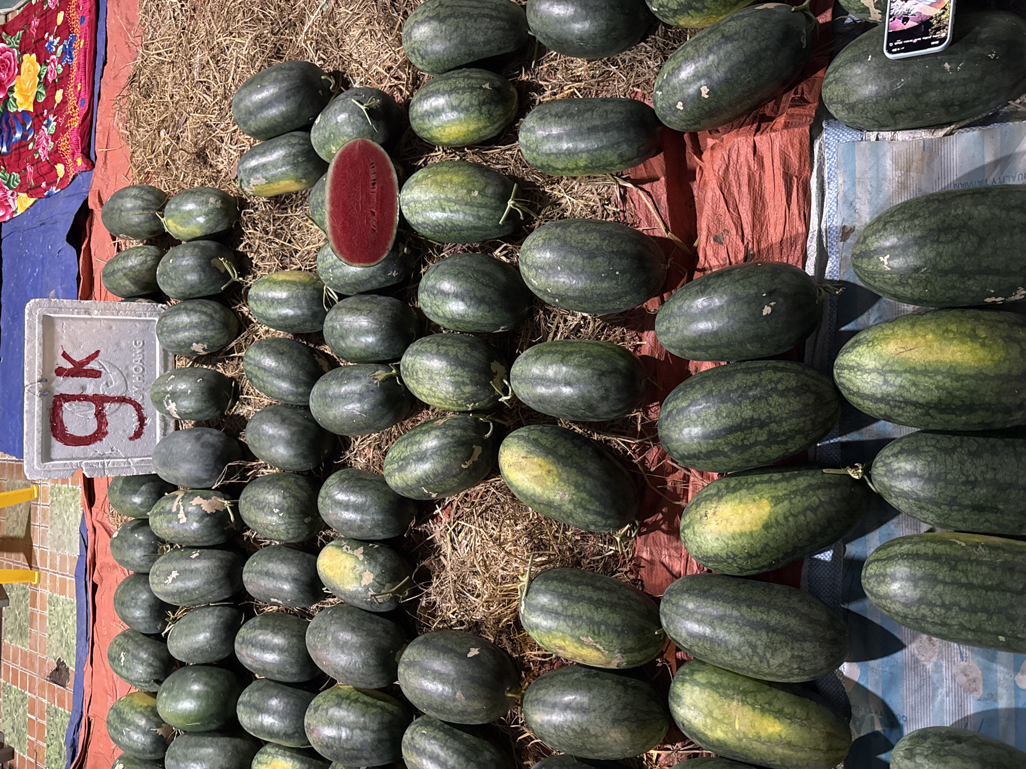 The image depicts a market stall in Đà Nẵng, Vietnam, overflowing with a plentiful harvest of watermelons. The fruits are arranged in various layers on a bed of straw and red tarp. Some watermelons are whole, while others are cut in half, revealing their vibrant red flesh and black seeds. A sign with the letters "gk" is visible in the background, likely indicating pricing or branding. The scene suggests a lively marketplace, possibly a local farmer's market or a roadside stand, where fresh produce is being sold. The lighting suggests it might be evening or under artificial light, giving a warm glow to the scene.
