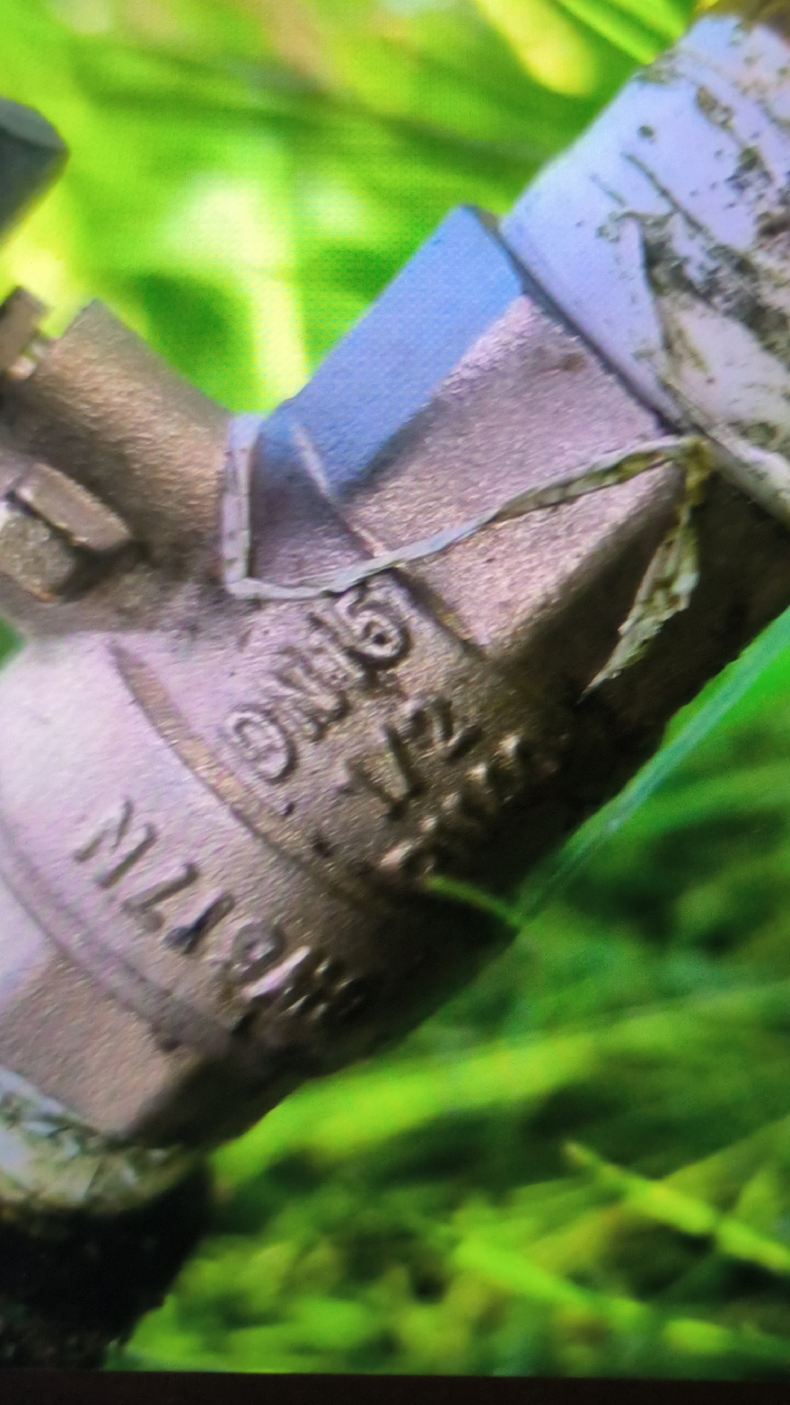 The image shows a close-up, angled view of a brass pipe fitting, possibly a valve, with embossed markings. The markings are partially visible and appear to include "DN15 1/2" and possibly other characters like "MZ1986". The fitting is metallic and shows signs of age or use. It is situated against a blurred background of vibrant green foliage, suggesting an outdoor or aquatic environment. There is no indication of the city of Kovrov, Russia, or any people in the image. The focus is entirely on the pipe fitting and its immediate surroundings.