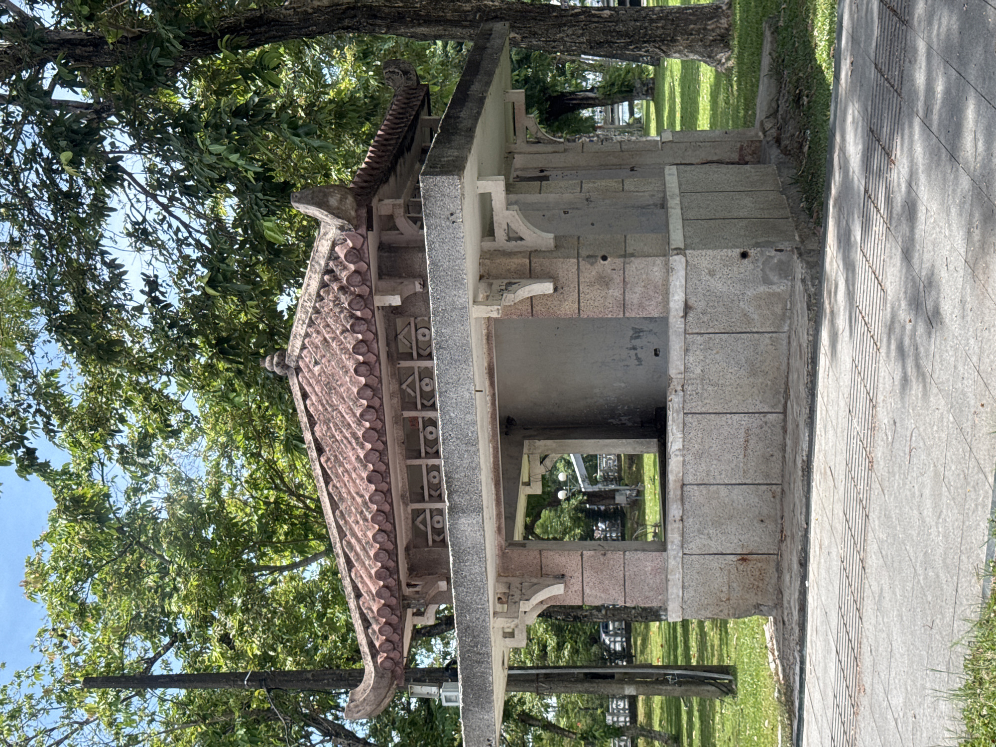 An ornate, abandoned kiosk with a traditional tiled roof stands in a park. The kiosk has a stone base and pillars, with a large open window. Green grass and trees surround the structure, and a paved walkway is visible in the foreground. The scene suggests a hot, sunny day. The kiosk is likely located in Phú Xuân Ward, Vietnam, given its architectural style.