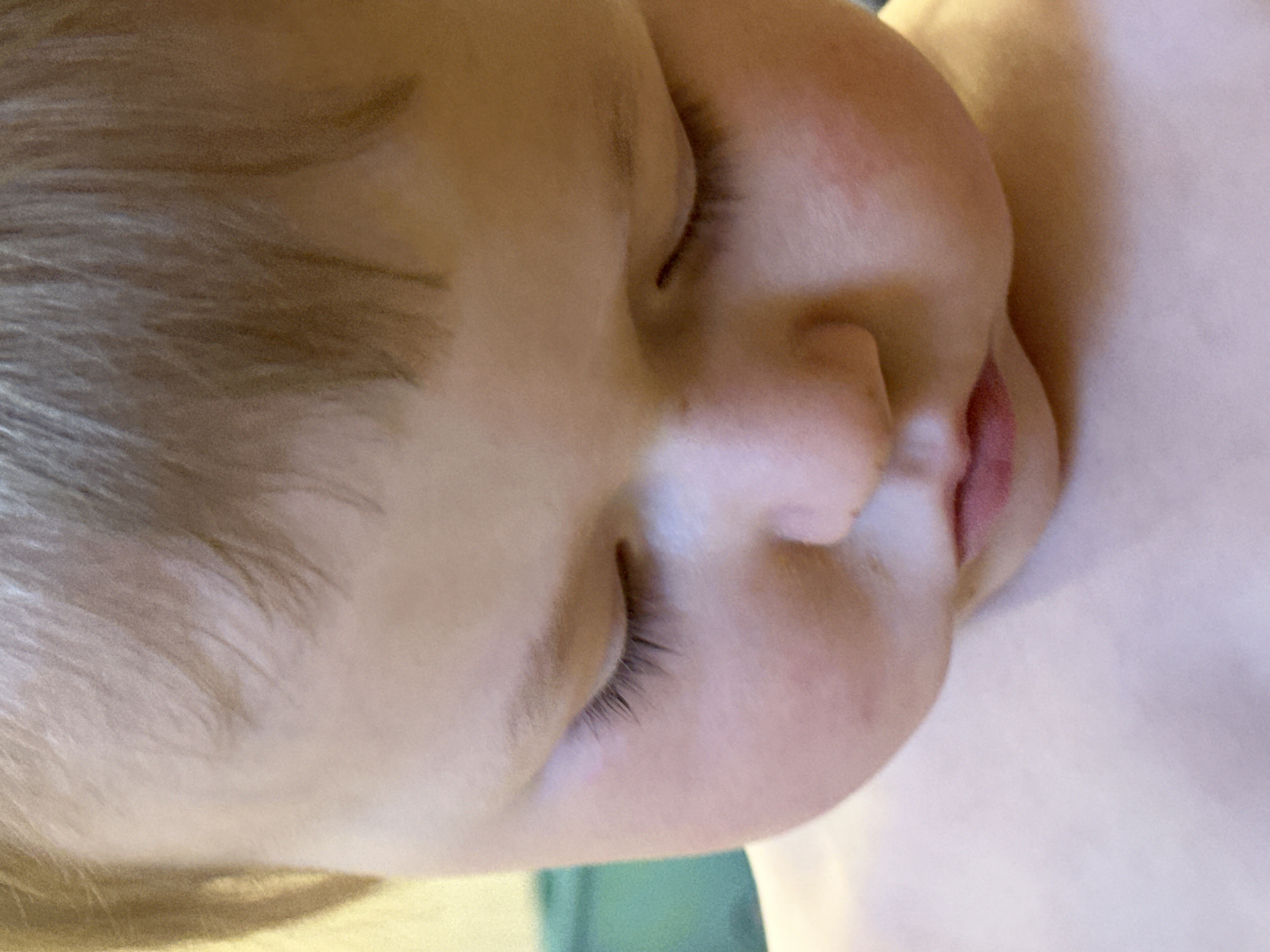 A close-up image depicts a young child with fair hair and closed eyes, presumably sleeping. The face is the main subject, showing the nose, mouth slightly open, eyelashes, and cheek. The skin tone is light. Parts of the child's shoulder and chest are visible. The background is indistinct but appears to be comprised of light and dark green hues. There is no indication of the location being Nizhny Novgorod, Russia.