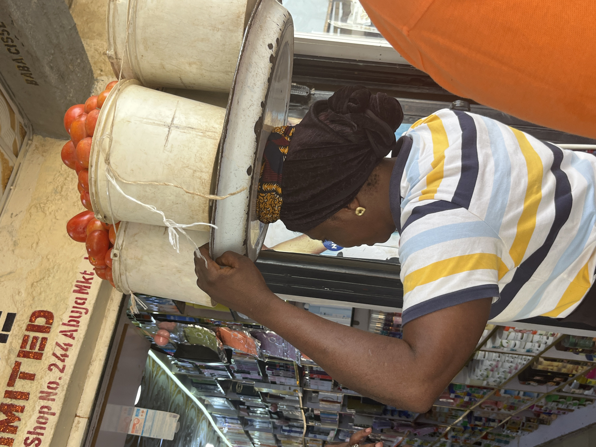 A person wearing a black headwrap and a striped t-shirt with yellow, blue, and white stripes is leaning over to tend to buckets of tomatoes. The buckets are stacked and appear to be made of a light-colored material. Red tomatoes are visible spilling out of one of the buckets. The person's left hand is reaching towards the bucket, and a gold earring is visible on their left earlobe. In the background, shelves filled with various items are visible, including what appear to be packaged goods. A sign on the left side of the image reads "LIMITED Shop No. 244 Abuja Mkt". An orange garment is draped over the top of the scene. The overall impression is of a market stall in Jos, Nigeria. The lighting suggests it is daytime.