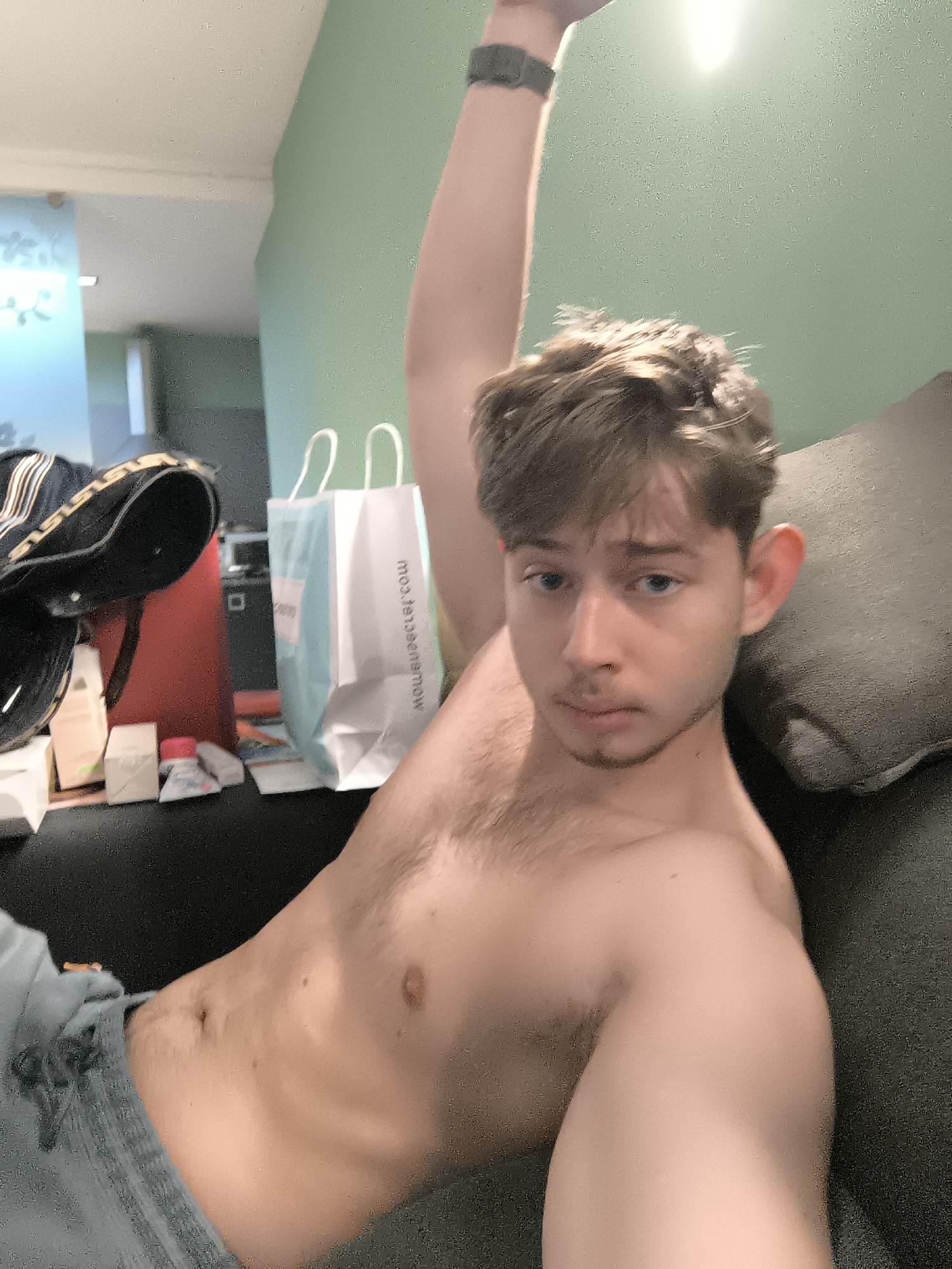 A shirtless young man is lying on a dark grey sofa. He has brown hair and blue eyes. His left arm is raised above his head. He is wearing grey jeans. In the background are various objects including a shopping bag, a red container, and a black bag. The background wall is light green. It is not possible to determine the location in Sofia, Bulgaria from this image.