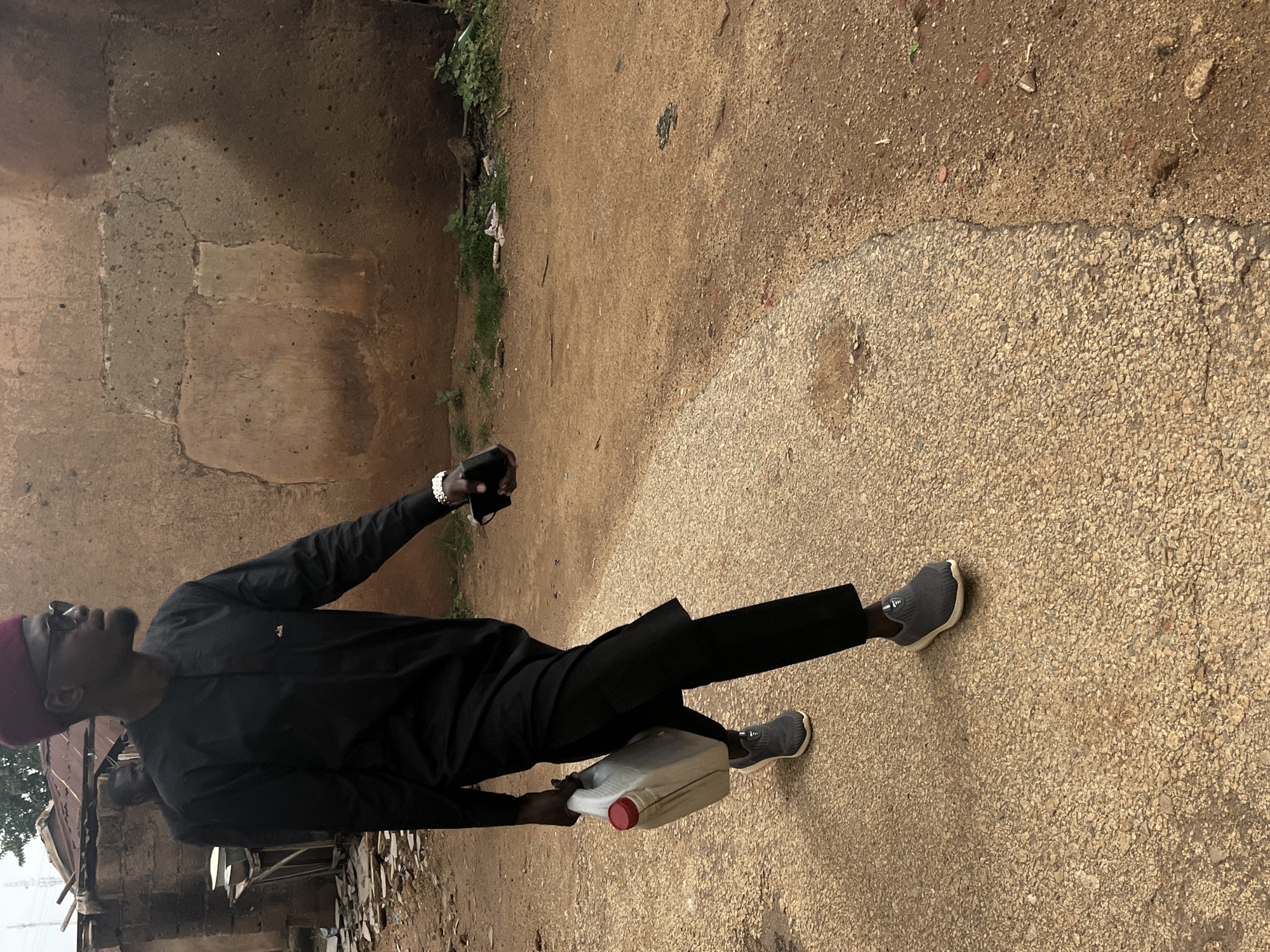 The image depicts a full-body shot of a Black man walking along a cracked asphalt path with a dirt shoulder in what appears to be a section of Jos, Nigeria, characterized by its rustic buildings and unpaved roads. The man, dressed in a black traditional outfit with a matching maroon cap, is seen from a slightly low angle. He carries a white jerrycan with a red cap in his left hand and a dark object, possibly a phone, in his right. His right foot is shod in a gray athletic shoe, while his left foot is also clad in a similar gray athletic shoe. The background reveals the textured, earthen walls of buildings, suggesting a warm, dry climate. A second individual, partially obscured, is visible behind the main subject, walking in the same direction. The environment is dusty and unkempt, with scattered debris and vegetation adding to the scene's authenticity. The lighting suggests it is daytime, with soft shadows.