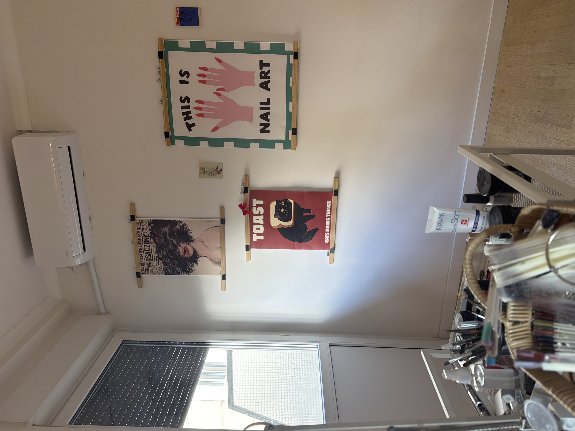 The image depicts an indoor space with several objects and decorations. On the wall are three posters: one with the text "THIS IS NAIL ART" and an illustration of hands; another with a cat sitting on a piece of toast, reading "TOAST: CATS DOING THINGS"; and a third with the text "TOM BERENGERI MIMI ROGERS" from the movie "OSAC ZONA". Above the "NAIL ART" poster is a small rectangular sticker that says "Silent Disco". To the left is a white air conditioning unit mounted on the wall.
Below the posters, a window with black blinds is visible. To the right is a cart filled with miscellaneous items including beauty and nail products like "EVELINE Extra Soft" lotion. The overall environment appears to be a room with a focus on nail art and cats, possibly in an apartment or studio. The location is not explicitly mentioned in the image.