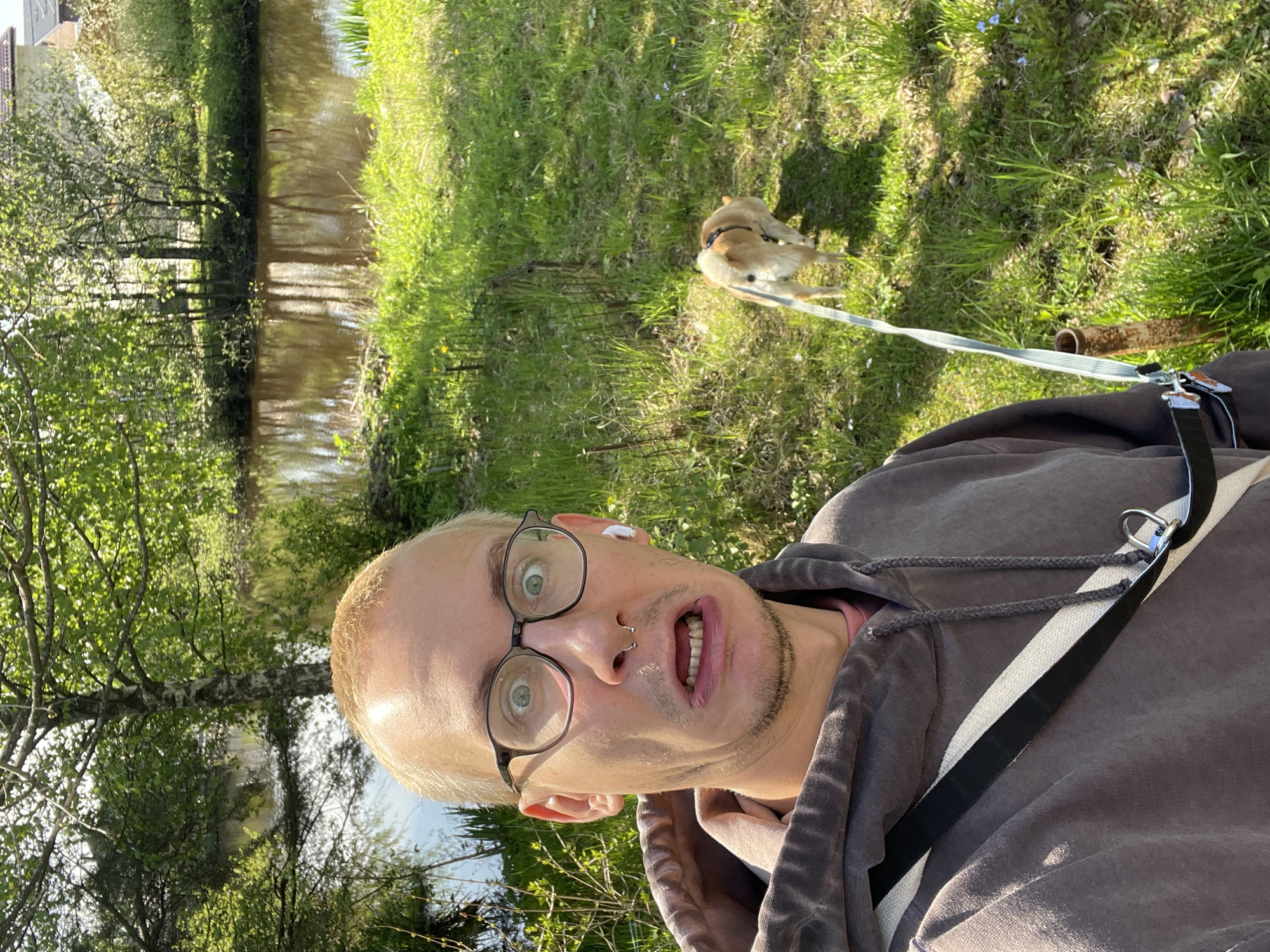 A light-skinned individual with fair hair and glasses is taking a selfie outdoors in Sestroretsk, Russia, adjacent to a canal or narrow river. The person is wearing a brown hoodie, and a strap or leash is visible, suggesting they are possibly walking a dog. A small brown and white dog is present on the right side of the frame, standing on a grassy area. The background shows green grass and trees, with the canal visible on the left side. The lighting suggests it is daytime.