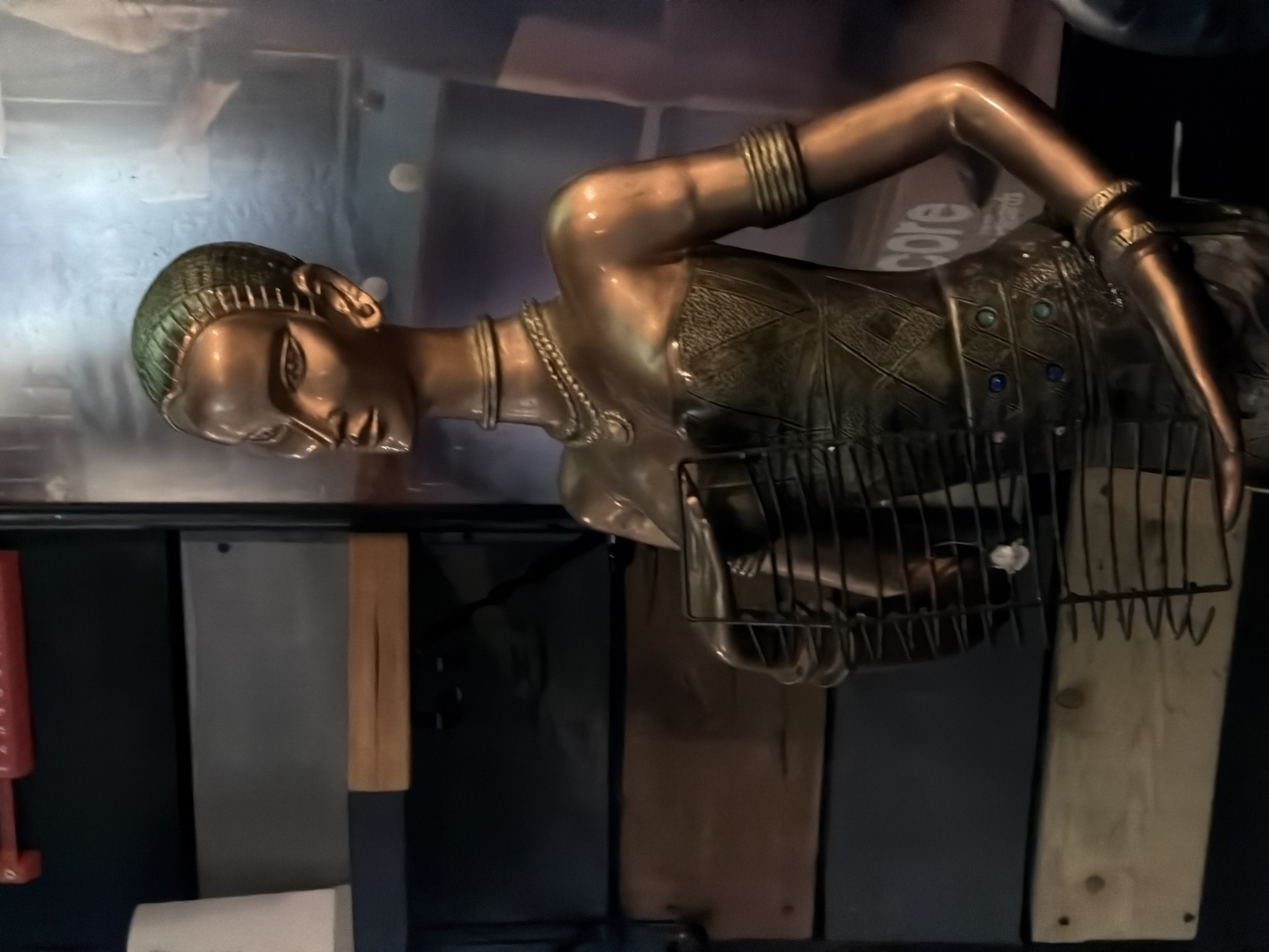 A bronze statue of an Egyptian-style woman reclines on a metal and wood surface in an indoor location. The statue is detailed with jewelry and patterned clothing, partly obscured by a metal rack. The background is blurred and suggests a kitchen or storage area with various tools and containers. The location in the city of Unknown, Unknown is not determinable from the image.