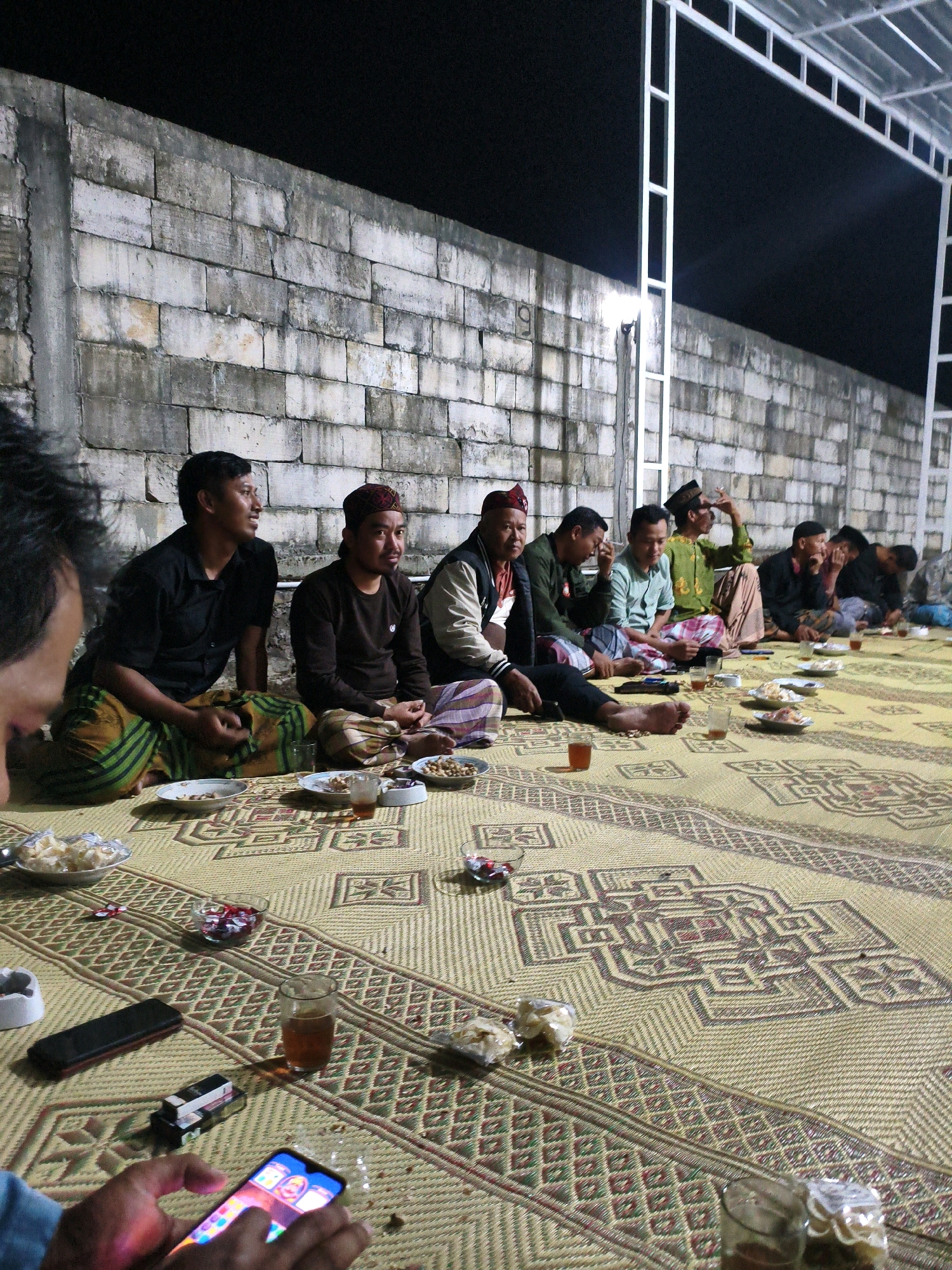 In Krajan Lor, Indonesia, a group of men are gathered at night, sitting on a woven mat on the ground. They are seated in a line against a textured, light-colored brick wall, with a white metal structure overhead suggesting an outdoor or semi-outdoor gathering space. The men are casually dressed, some wearing traditional Indonesian attire such as sarongs and peci (caps). On the mat in front of them are various snacks and drinks served in small bowls and glasses, indicating a communal meal or gathering. Some men are engaged in conversation, while others are looking down, possibly at their phones or the food. The lighting is dim, with artificial light illuminating the scene from above and behind the camera, casting soft shadows. One man in the foreground is looking at his smartphone, which is displaying colorful icons. The overall atmosphere appears relaxed and social.