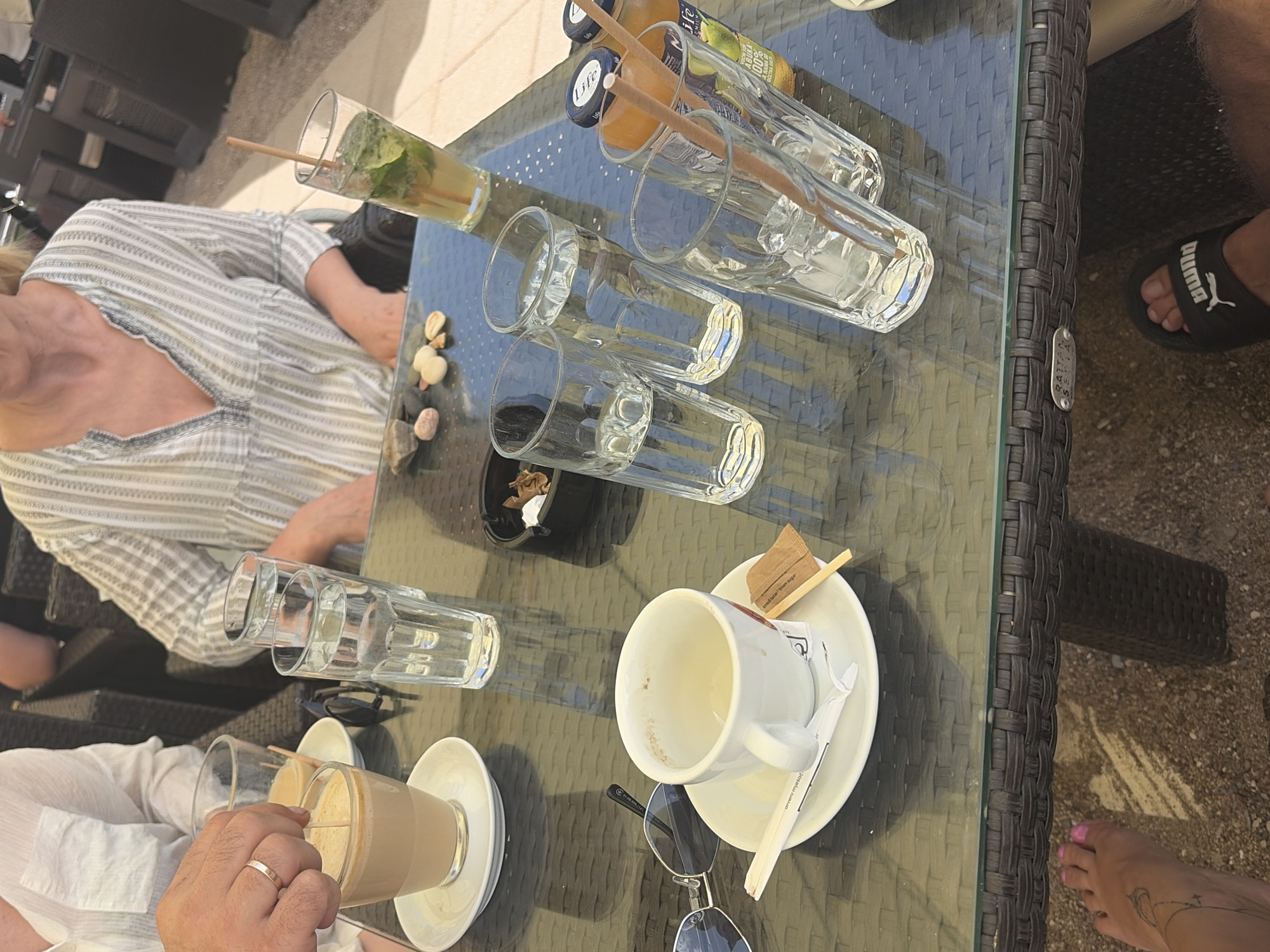The image depicts a table setting at an outdoor cafe, likely in Budva, Montenegro, based on the details of the furniture. The table, made of dark woven material with a glass top, is laden with several drinks and items. In the foreground, a white ceramic cup and saucer sit with a partially emptied beverage and used sugar packets. Nearby, a pair of sunglasses rests on the table. A hand, wearing a ring, is visible stirring a frothy drink in a glass.
Several other glasses are present, some containing clear liquids and others with what appears to be a mojito or similar cocktail garnished with mint. A bottle of juice or iced tea with a "Lipa" label is also on the table. The background shows the lower torsos of two individuals seated at the table, one wearing a striped top and the other a white shirt. The ground beneath the table appears to be sand or gravel, characteristic of a beachside or coastal establishment. The overall scene suggests a casual, leisurely moment at an outdoor cafe.