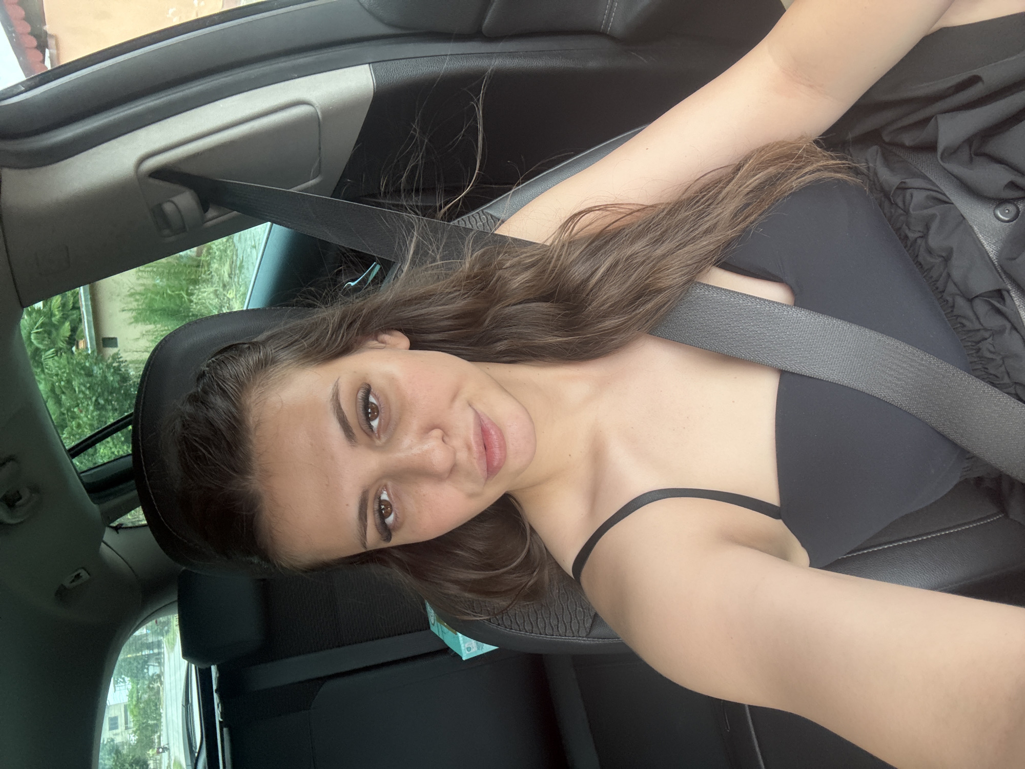 In Govedare, Bulgaria, a young woman with long, dark brown hair is pictured inside a vehicle, seated in the driver's seat. She is wearing a black top and a grey seatbelt across her chest. The interior of the car is visible, including the black leather seats and the car's pillar. Through the car window, a rural outdoor environment can be seen, with trees and buildings in the background. The overall lighting suggests it is daytime.