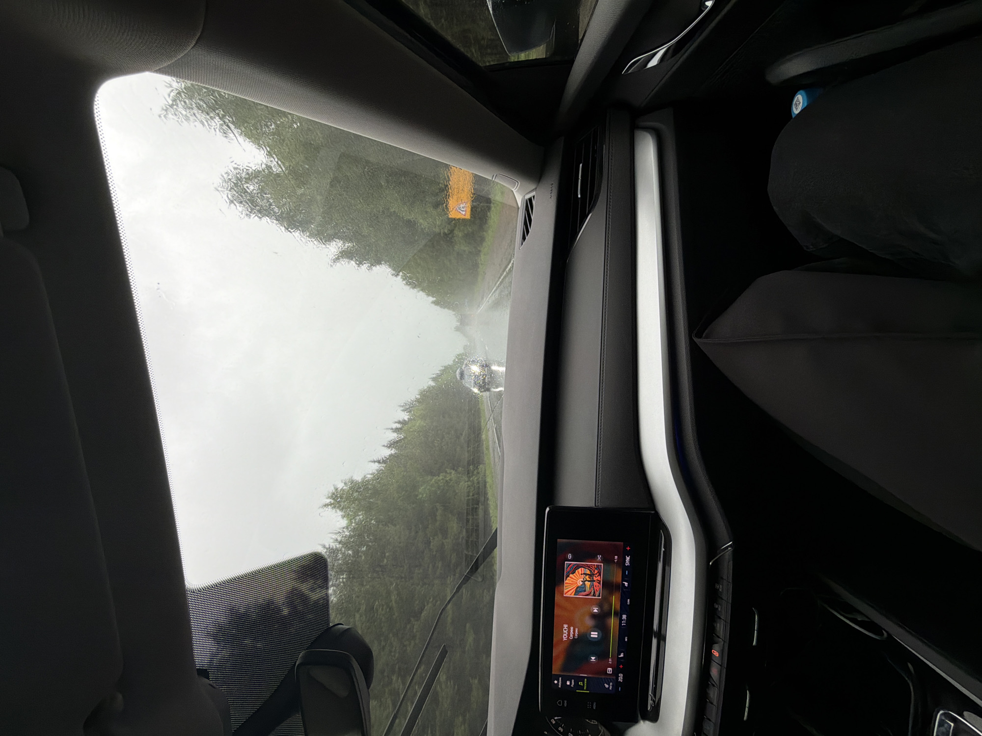 The image shows the view from inside a car driving on a road in Dvigatel', Russia. It is raining heavily, and the windshield wipers are on. There is a vehicle ahead on the road. The car's infotainment screen is visible, displaying music information. A road sign is visible, blurry in the distance. A passenger seat is present in the frame but not occupied. The overall scene suggests travel in inclement weather.