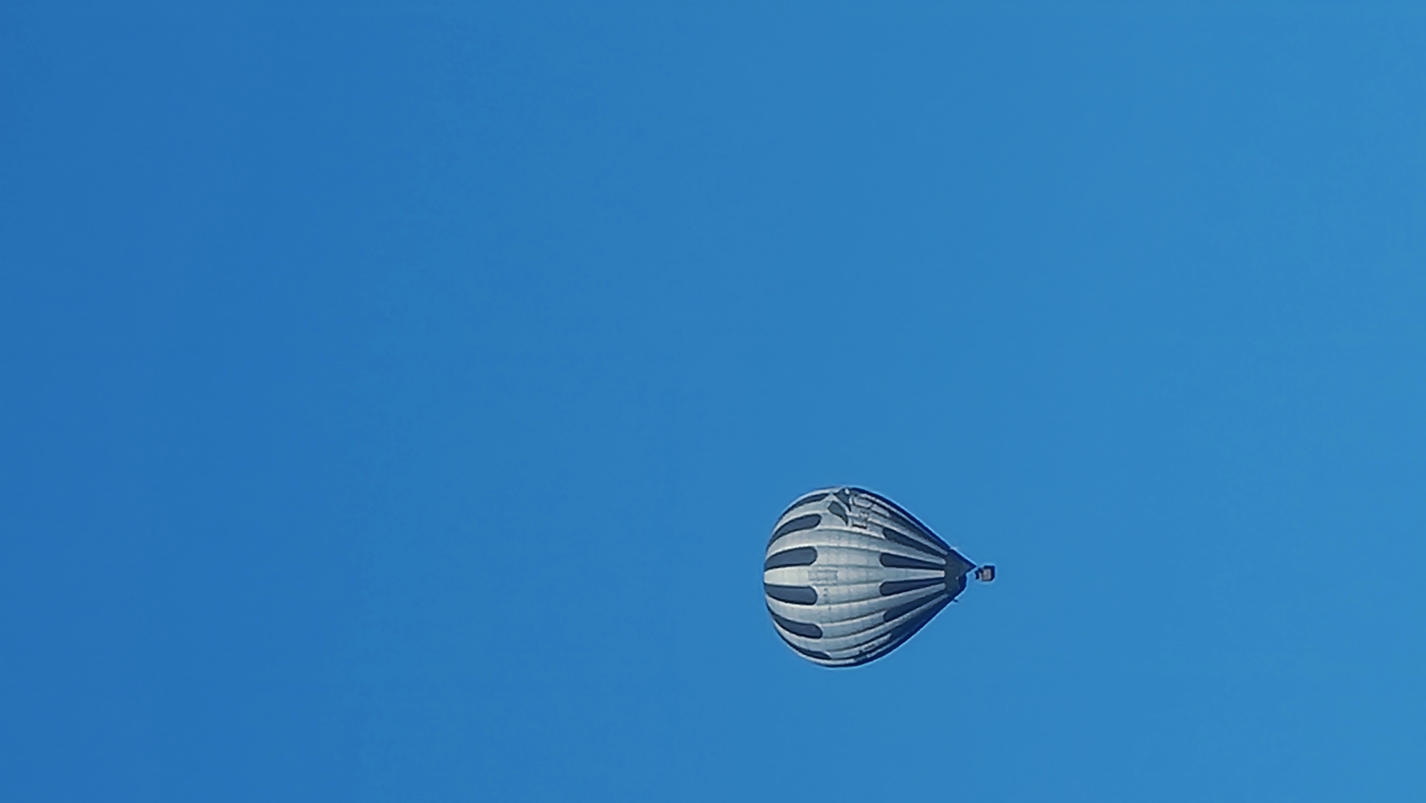 A hot air balloon with a black and white striped pattern is floating in a clear blue sky. The balloon is in the lower left quadrant of the image. A small basket is suspended beneath the balloon. The background is entirely sky, with no other objects or landscape visible. The scene is devoid of human activity and appears to be taken from ground level looking up. There is no discernible location in the city of Sant Joan Les Fonts, Spain.