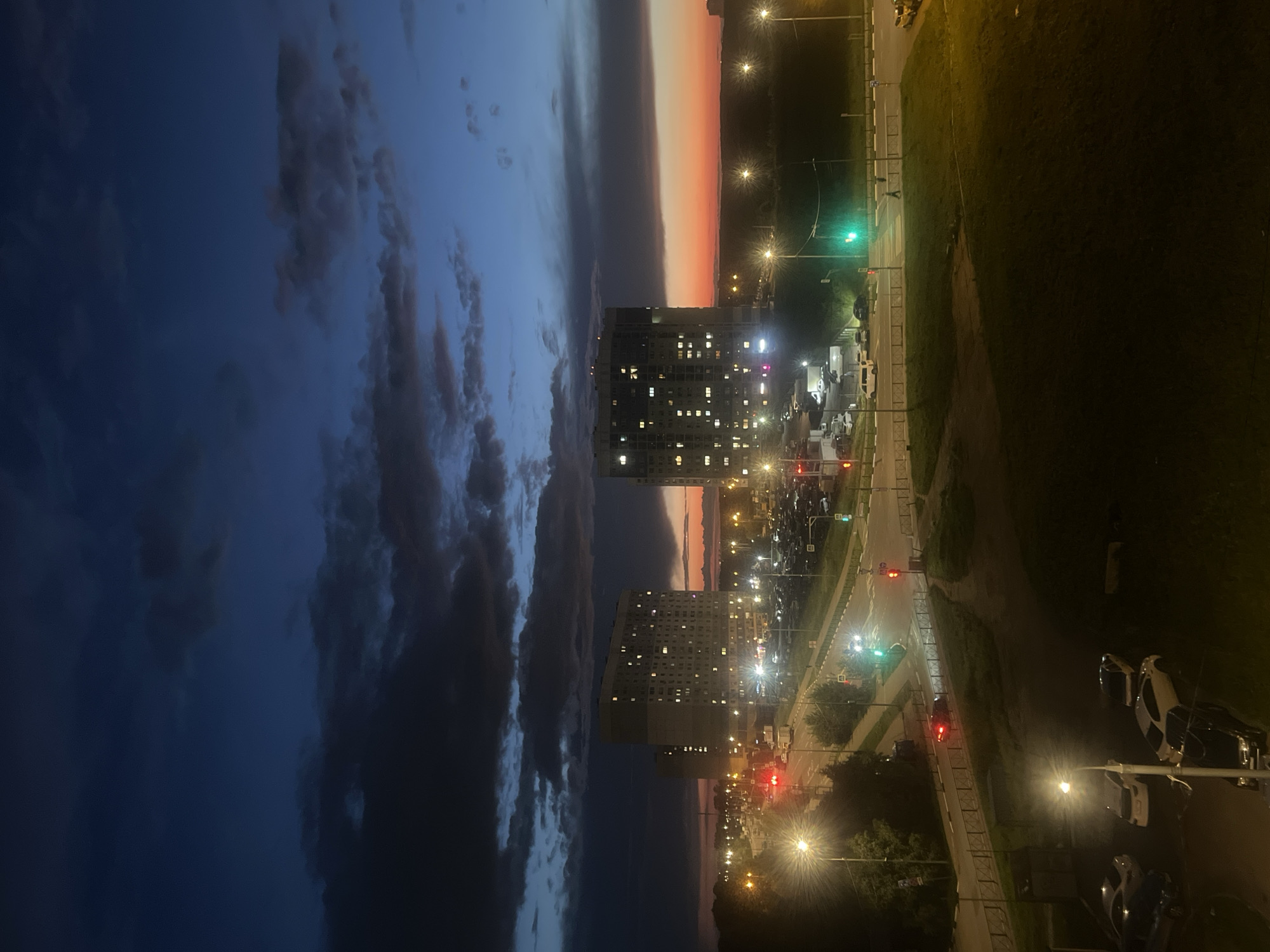 The image depicts a twilight scene over a residential area in Domodedovo, Russia. The sky transitions from a deep blue on the left, filled with dramatic, dark clouds, to a warm gradient of orange and pink hues near the horizon on the right, suggesting the end of the day. Two tall, modern apartment buildings with illuminated windows are prominent features.
Below the buildings, a street is visible, lined with streetlights casting a yellow glow. Several cars are parked along the street and in a designated parking lot. Traffic lights are also visible, one showing green and others red. The scene includes patches of grass and trees, indicating an urban or suburban parkland. The overall mood is peaceful, with the artificial lights of the city contrasting with the fading natural light of the sky. The orientation of the image is vertical. No visible text is present in the image.