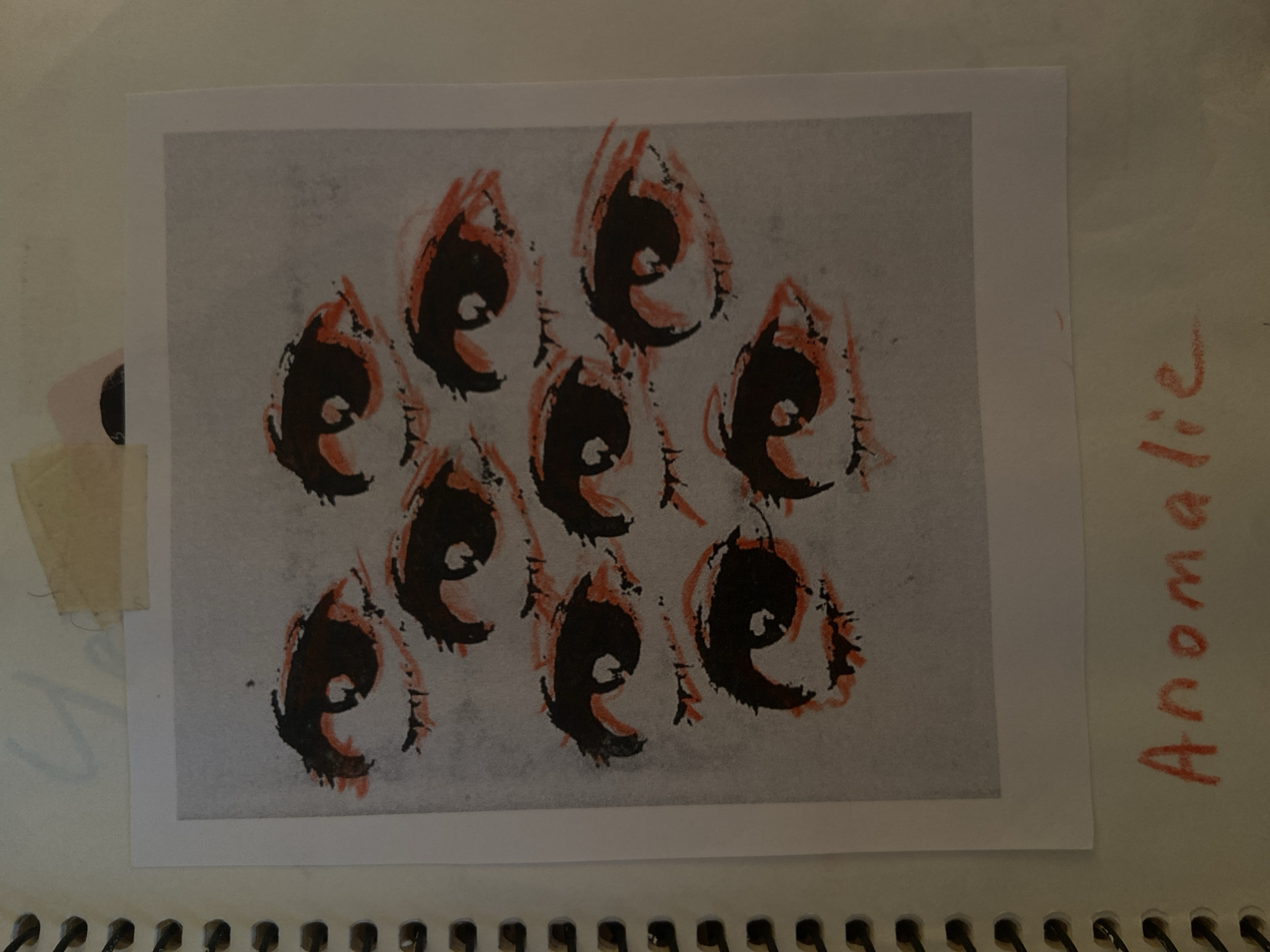 The image shows a close-up, overhead view of a spiral-bound notebook page. The notebook has a white cover with a gray spiral binding visible on the left side. A white piece of paper, appearing to be a photograph or print, is taped to the notebook page with a piece of light brown tape at the top. This paper features a drawing of multiple pairs of eyes with dark pupils and reddish-orange outlines around the eyelids. Below the drawing, the word "Anomalie" is written in red crayon on the notebook page. Faint blue handwriting is visible on the notebook page above the taped drawing. The setting is an indoor environment, likely a desk or table, with neutral lighting. No specific location in Krasnodar, Russia can be identified from this image.