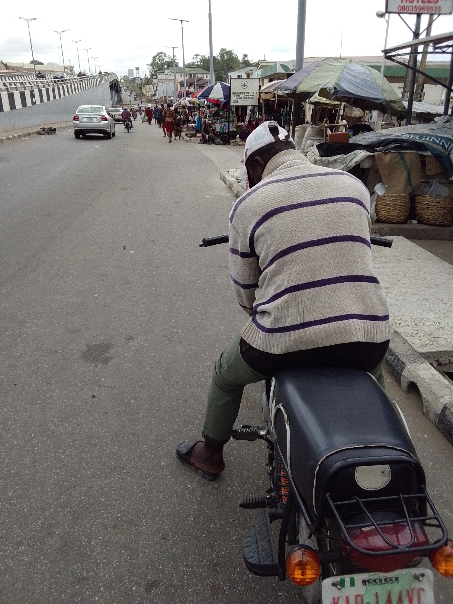 The image shows a street scene in Lokoja, Nigeria. A man wearing a striped sweater, dark pants, and a white cap is sitting on a motorcycle facing away from the camera. He is wearing slide sandals. The motorcycle is parked near the edge of the road. In the background, there is a bridge, a silver car, and people walking on the sidewalk and in the street. There are also market stalls with umbrellas and goods for sale. A sign for "CHIEF MAGISTRATE COURT II & III LOKOJA JUDICIAL DIVISION, LOKOJA, KOGI STATE" is visible, which helps identify the location. There are several pedestrians in the background. A sign that reads "HOTELS" is also visible.