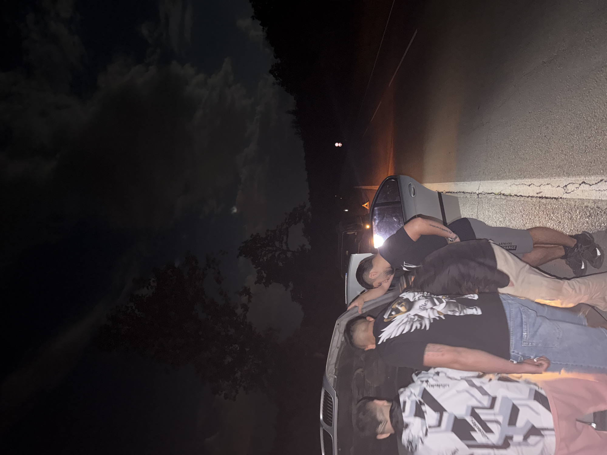 At night in an unknown city, several individuals are gathered near a silver-colored car parked on the side of a road. The sky is cloudy. Three men are standing by the car, one wearing a black shirt with a white eagle design and blue jeans, another a black shirt with "ARMANI EXCHANGE" visible on grey shorts, and the third in a black and white patterned shirt. One of the men appears to be looking under the hood of the car. The road surface is paved and lined with white paint. Headlights are visible in the distance.