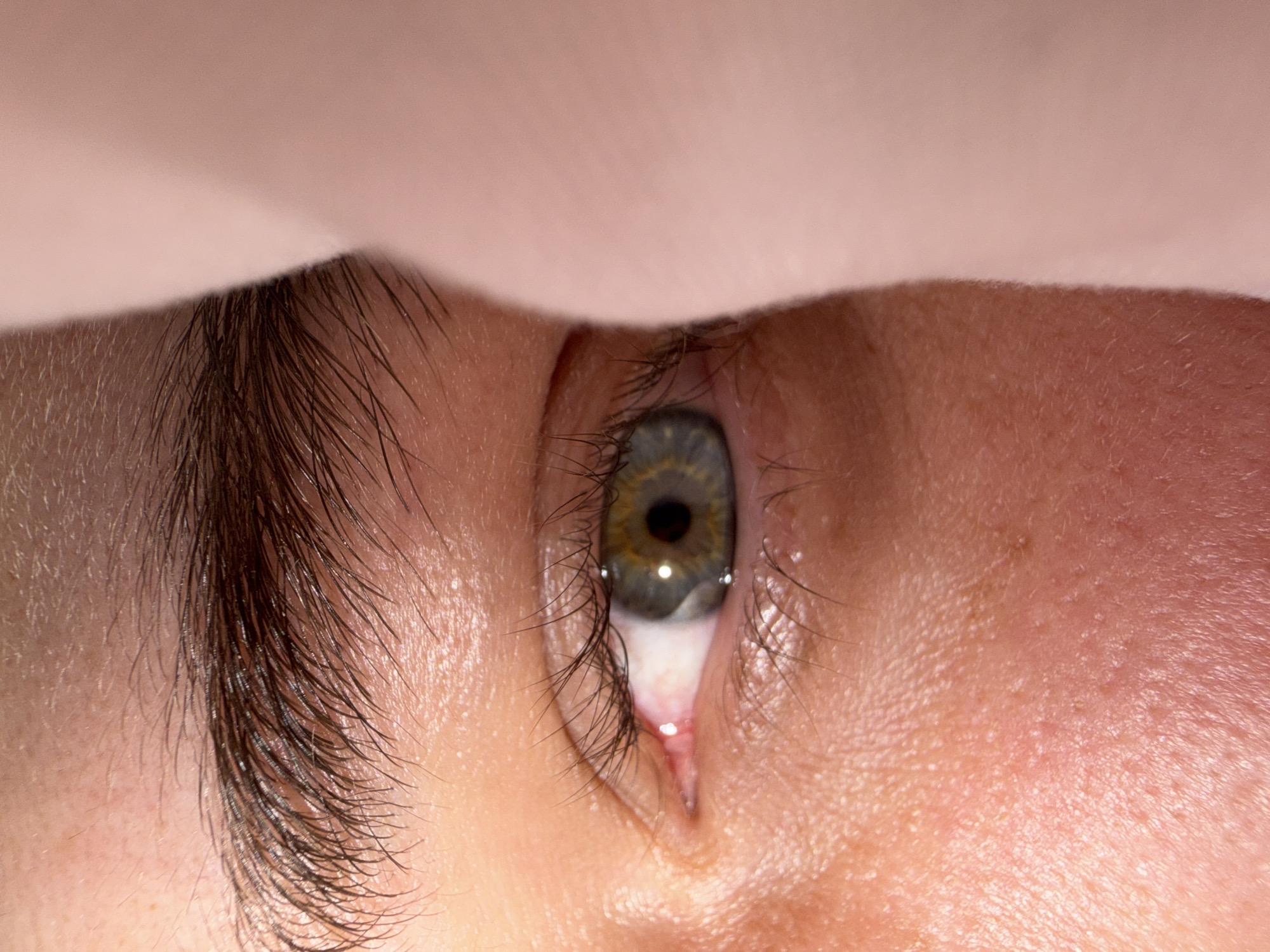 This is a close-up, macro photograph of a person's eye, seen from a low-angle perspective. The eye is the central focus, with the iris exhibiting a multi-colored pattern of green, gold, and grey, surrounding a dark pupil. Eyelashes frame the eye, and the lower eyelid is slightly retracted, revealing the pink inner membrane. The surrounding skin appears fair and is textured with pores. Above the eye, a soft, peachy-colored material is draped, casting a shadow and obscuring the upper part of the subject's face, suggesting a person is looking upwards or is partially covered. The lighting is direct, creating a slight glare on the eye's surface. There is no visible text in the image. The setting context is Bangkok, Thailand.