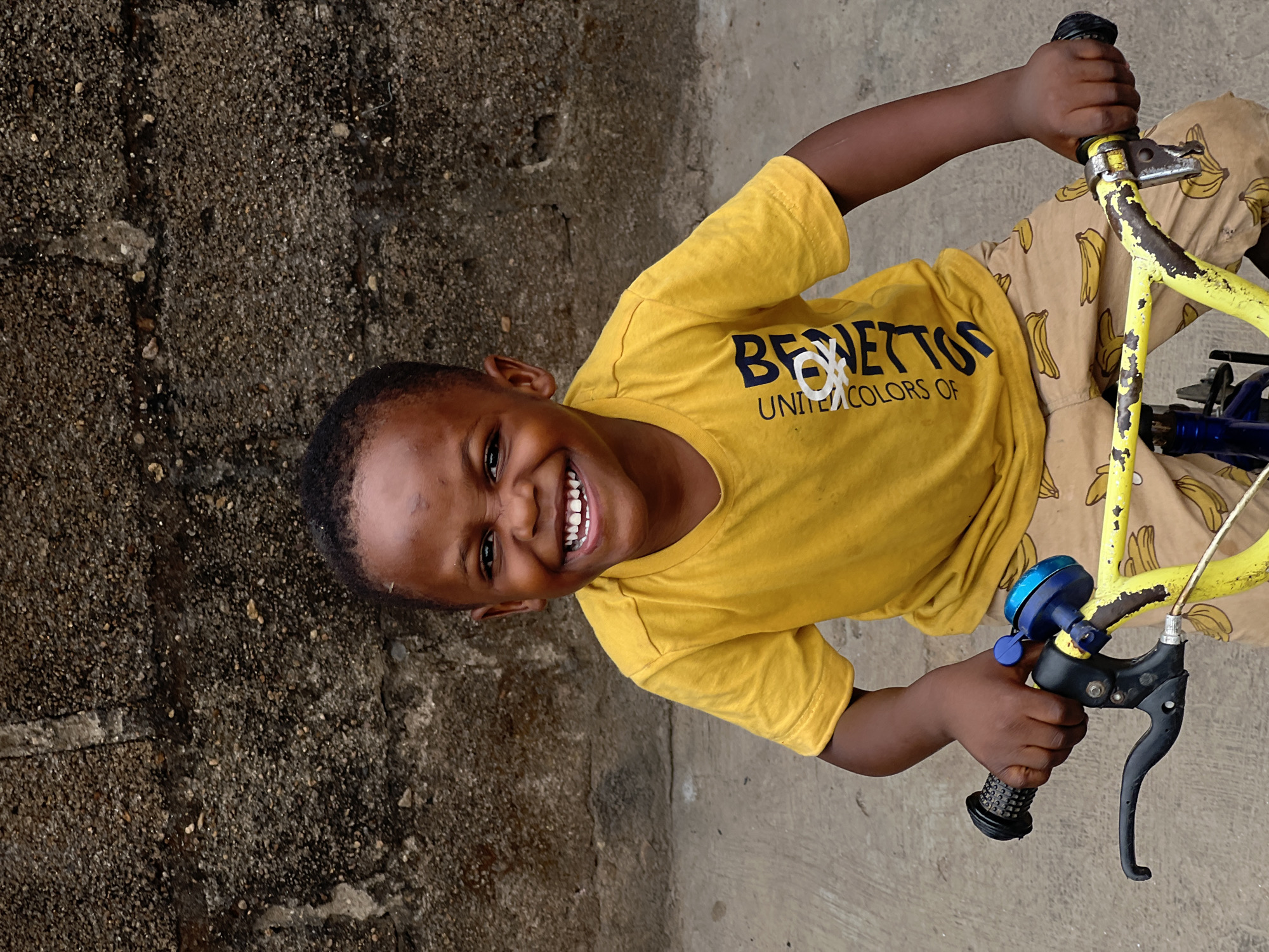 A vibrant, vertically oriented full-shot image captures a young Black child, likely a boy, joyfully posing on a bicycle outdoors in Jos, Nigeria. The child, with short dark hair, is smiling broadly at the camera, revealing white teeth, and conveys an expression of happiness.
He is wearing a bright yellow short-sleeved t-shirt with the text "BENETTON" and "UNITED COLORS OF" printed in dark blue across the chest, partially covered by a white circular logo. His pants are a light brown or tan color, adorned with a repeating pattern of yellow bananas.
The child is seated on or straddling a bicycle with a distinctive yellow frame that shows signs of wear, with chipped paint revealing darker material underneath. His hands firmly grasp the black handlebars, and a blue bicycle bell is visible on the left side, along with a black brake lever.
The background features a textured, dark gray or brown wall on the left, possibly concrete or stucco, and a lighter, smoother concrete or asphalt ground surface on the right. The bright lighting indicates it is daytime. The image captures a moment of cheerful innocence in an outdoor setting.