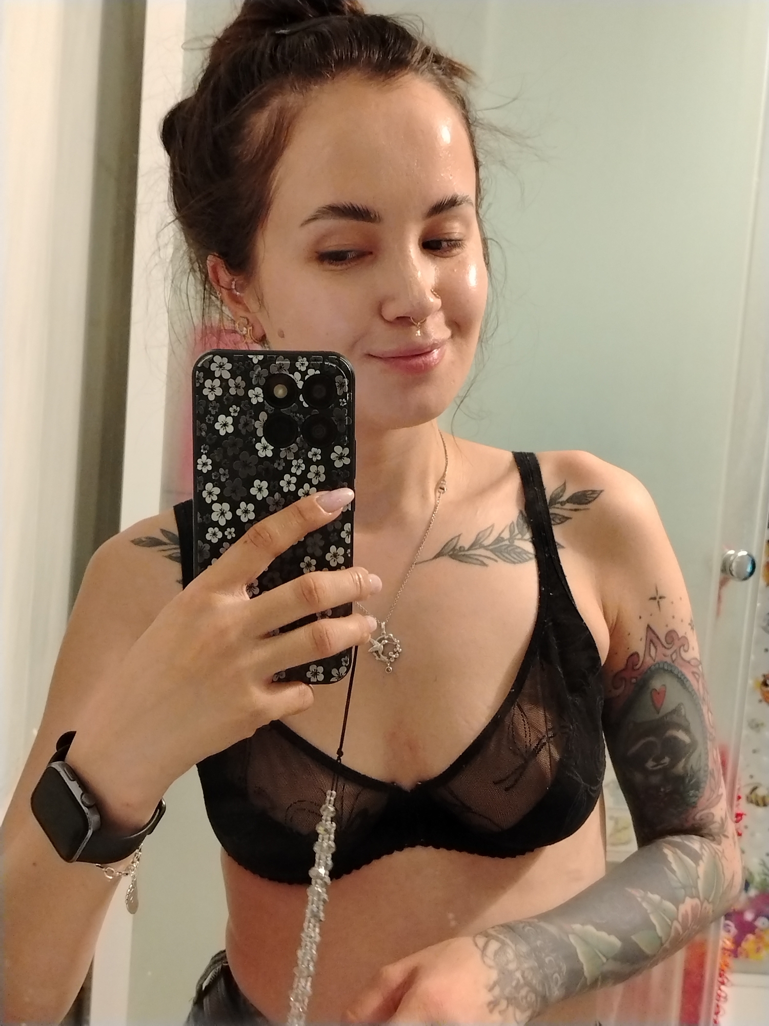 A woman is taking a selfie in a bathroom. She has her hair in a messy bun and is wearing a black, sheer bra. She has multiple tattoos on her arms and chest, including a raccoon tattoo on her right arm. She is holding a smartphone with a floral case. She is also wearing a smartwatch on her left wrist. The background is a bathroom with a mirror and a white or light green wall. There is no indication of a location in the city of Noviy, Russia.
