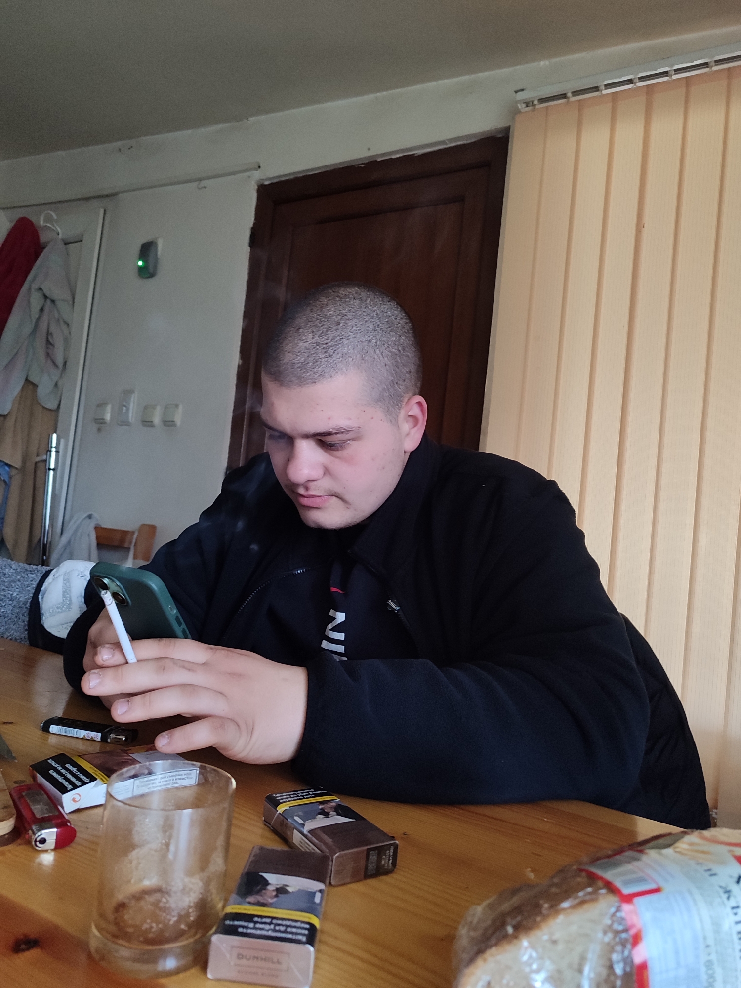 A young man with a shaved head is seated at a wooden table, holding a lit cigarette in his left hand and a green smartphone in his right. He is wearing a black zip-up jacket over a shirt with "IN" printed on it. On the table, several packs of Dunhill cigarettes, two lighters, a dirty glass, and a loaf of bread in plastic packaging are scattered. In the background, a white wall with a green device, a partially visible wooden door, and beige vertical blinds are seen. A garment hangs on a door in the distant background. The scene gives the impression of an interior setting in Bulgaria; the OCR shows Cyrillic text in the cigarette packaging. There is no mention of the city of Varshilo.