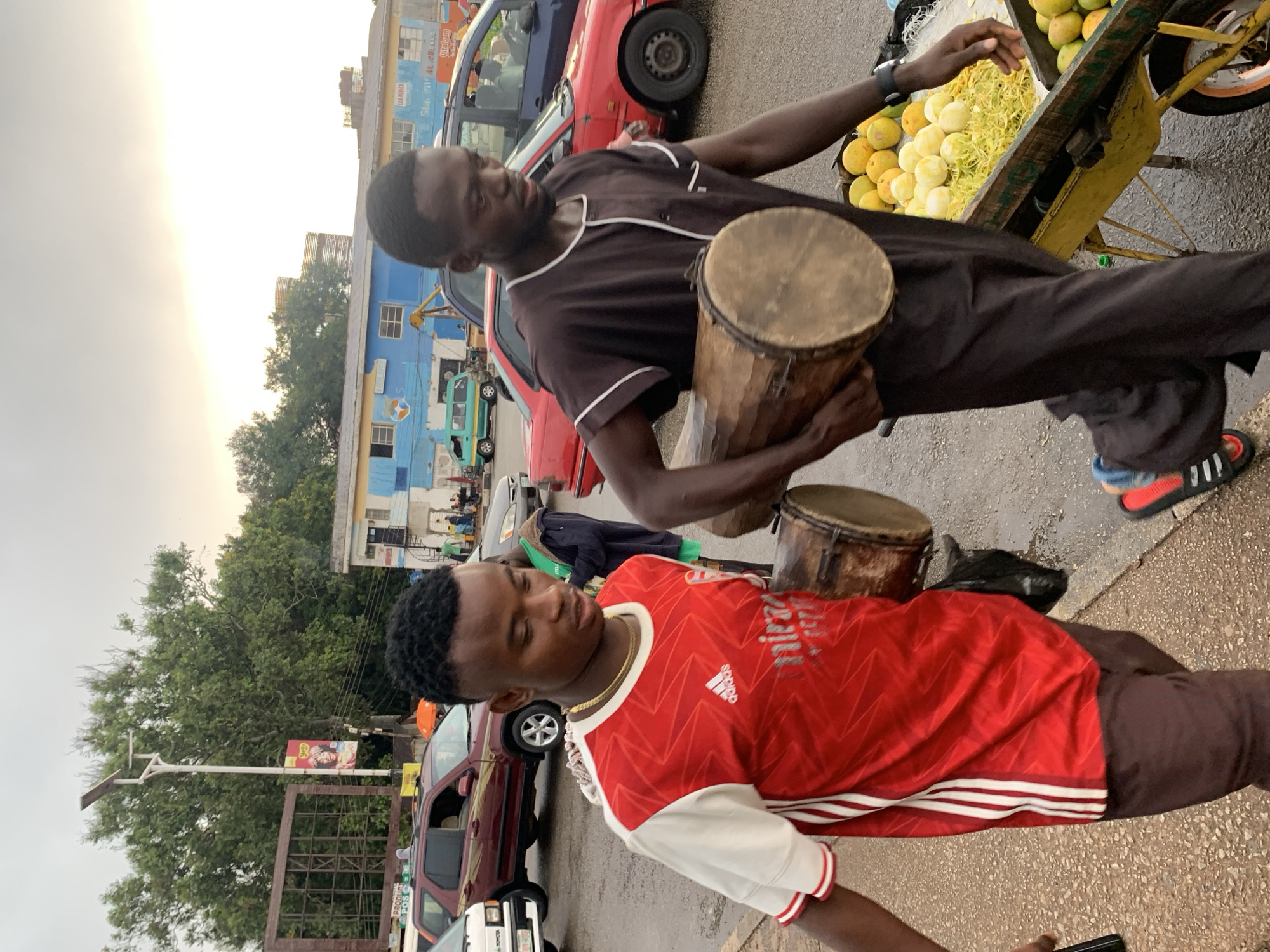 Two young Black men are walking on a wet, paved street in Jos, Nigeria, during the daytime under an overcast sky. The man on the left is wearing a red and white short-sleeved Adidas jersey with white stripes on the shoulders and brown pants. He is looking down and to the right. The man on the right is wearing a dark brown short-sleeved tunic with white piping and dark pants. He is carrying a large, cylindrical wooden drum horizontally with his right arm. In front of him is a yellow cart loaded with fruits, including what appear to be oranges and shredded yellow produce. The background shows a busy street with several vehicles, including a red SUV, a red sedan, and a blue car. Buildings line the street, some of which appear to be shops with signage. A lamppost with a solar panel stands near a large tree. The overall impression is a candid street scene capturing everyday life and commerce in a Nigerian city.