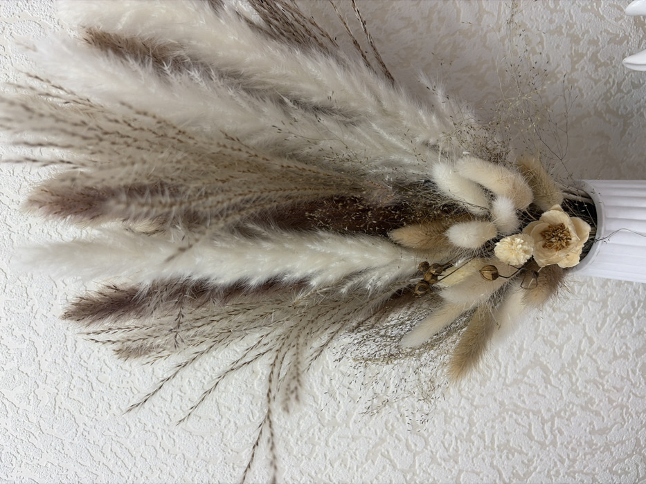 The image displays a detailed close-up of a decorative arrangement primarily composed of dried natural elements, set against a textured white wall. The arrangement features a variety of dried grasses and flowers, presented in a fan-like or cascading style.
The most prominent components are large, fluffy plumes, likely pampas grass, which range in color from creamy white to light beige and a deeper, muted brown or purplish-brown. These plumes have a soft, feathery texture and are arranged to spread out towards the left side of the frame. Mixed in with these larger elements are finer, wispy dried grasses with delicate seed heads, adding subtle texture and depth. These finer grasses are also in neutral shades of light brown and beige.
Towards the right side of the arrangement, a cluster of smaller, softer, and more rounded fluffy elements, possibly dried bunny tail grass (Lagurus ovatus), is visible in shades of light beige and warm brown. Within this cluster are a few dried floral accents: a light cream or pale yellow flower with a distinct, slightly darker brown center, and a smaller, cream-colored, fluffy dried flower next to it. Additionally, a few dark, spherical seed pods or berries on thin stems add a touch of darker contrast.
The arrangement is held within a white, ribbed or fluted vase, a section of which is visible on the far right edge of the image. The background is a white or off-white wall with a visible, rough stucco or textured wallpaper pattern, providing a subtle, complementary backdrop. The lighting is soft and even, emphasizing the varied textures and natural color palette of the dried botanicals. The overall aesthetic is natural, rustic, and decorative. The image appears to be rotated 90 degrees clockwise from its likely upright display orientation.