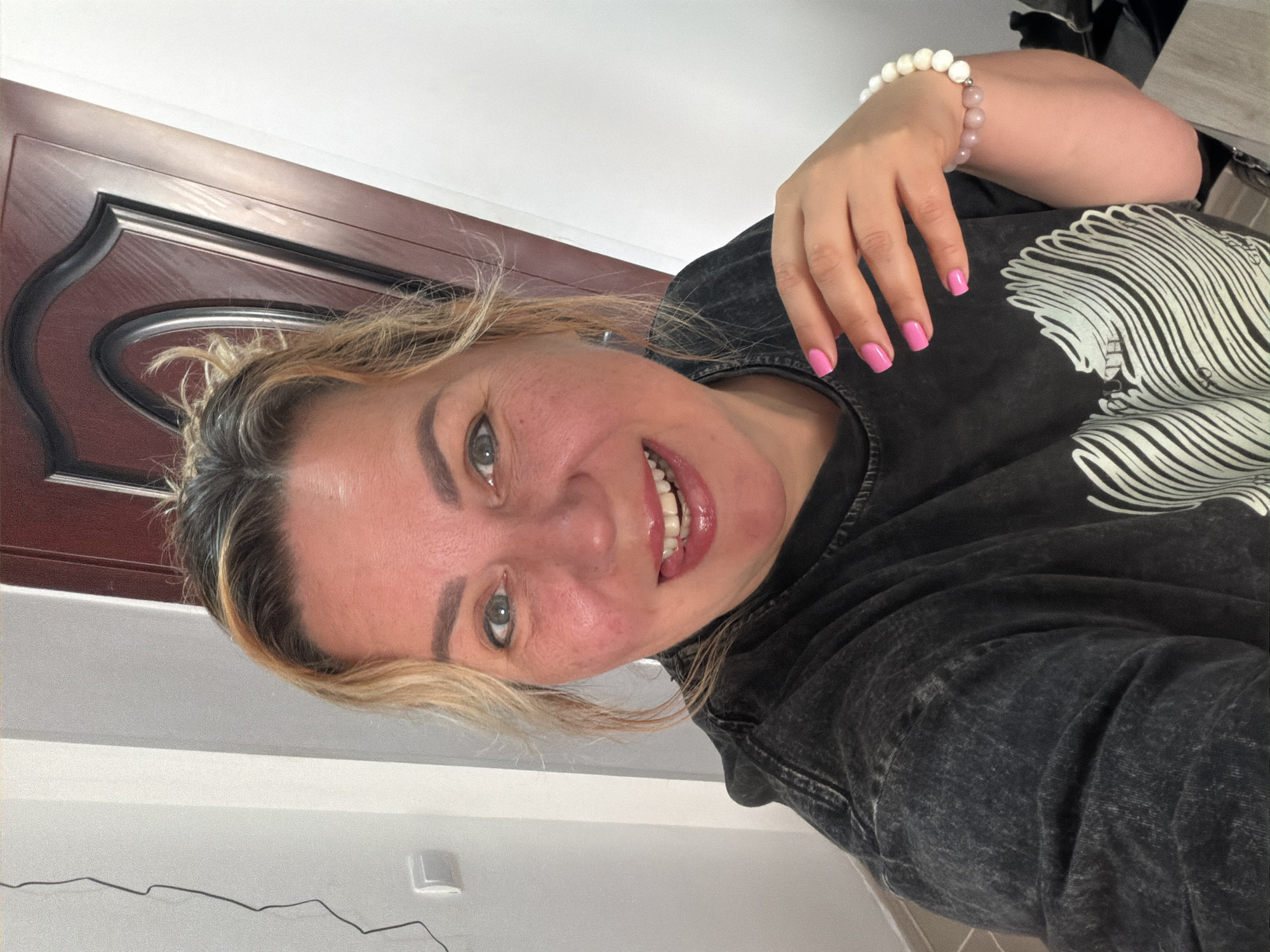 A woman with blonde hair pulled back in a bun is smiling towards the camera. She is wearing a dark, distressed T-shirt with a white design on the front. She has a bracelet made of white and pink beads on her left wrist. Her fingernails are painted pink. Behind her is a brown door with a decorative trim and a white wall. The location is Leningradskaya, Russia.