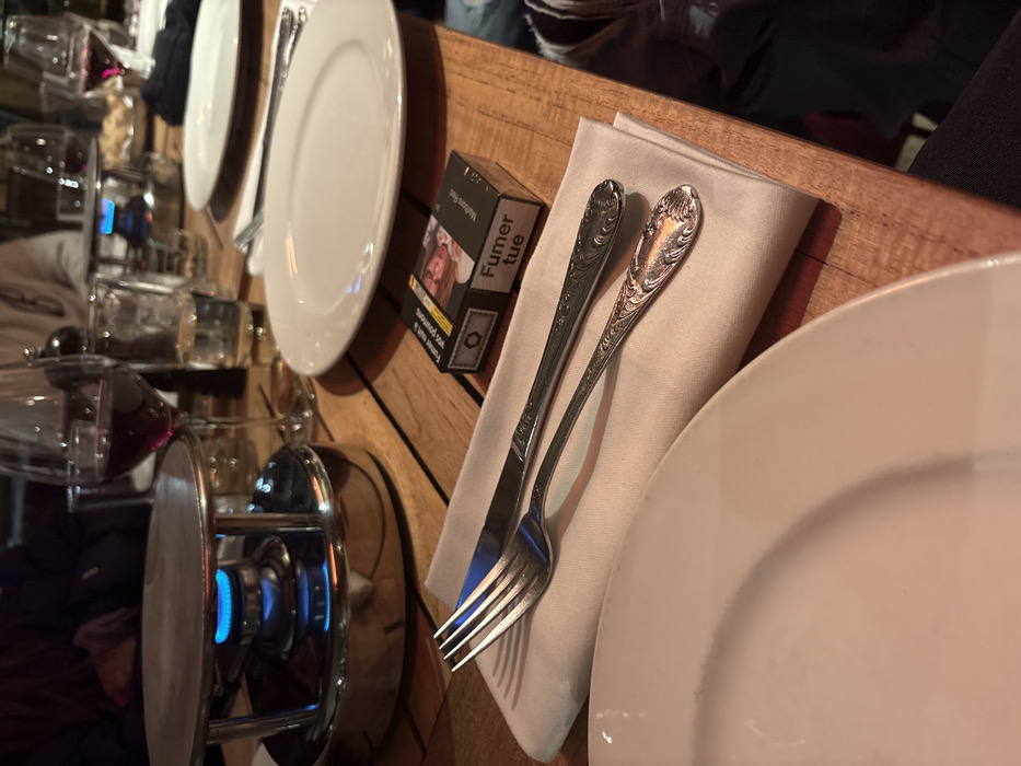 The image depicts a dining table scene in a restaurant in La Clusaz, France. The foreground features a partial view of a white ceramic plate. To its right, a cream-colored cloth napkin is placed, holding a fork and a knife with ornate handles. Adjacent to the napkin, a black pack of Marlboro Red cigarettes lies on the wooden table, displaying the French health warning "Fumer tue" (Smoking kills) and a graphic image.
In the mid-ground, a stainless steel fondue burner with a blue flame is active, positioned beneath a flat metal surface. Behind this, a wine glass filled with red wine and a clear glass of water are visible. Further back, another white ceramic plate is set, accompanied by a fork and another wine glass containing red wine. The background is blurred but shows indistinct figures of people seated at the table, suggesting a social dining environment. The table surface is constructed from light-colored wood planks.