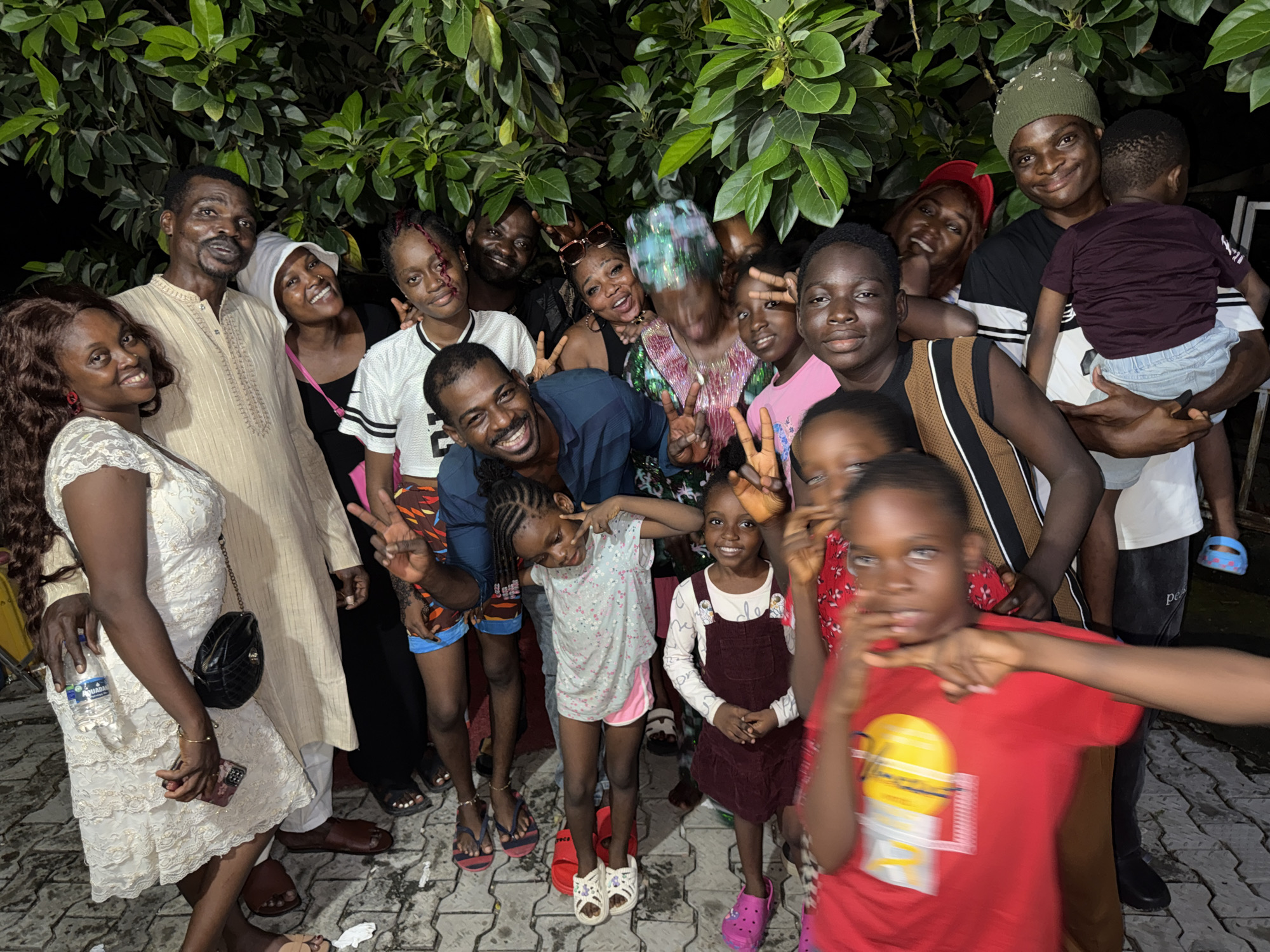 A group of approximately 16 individuals, including adults and children, are posed together for a photograph against a backdrop of lush green foliage, possibly within the city of Ado, Nigeria. The setting seems to be an outdoor gathering. Individuals are dressed in casual attire. Several people are smiling and making peace signs with their fingers. A woman on the left wears a white dress and holds a black purse. A man next to her is wearing a cream-colored tunic. A man on the right holds a young child. The ground appears to be paved with interlocking concrete pavers. A water bottle and a cell phone are also visible in the image.