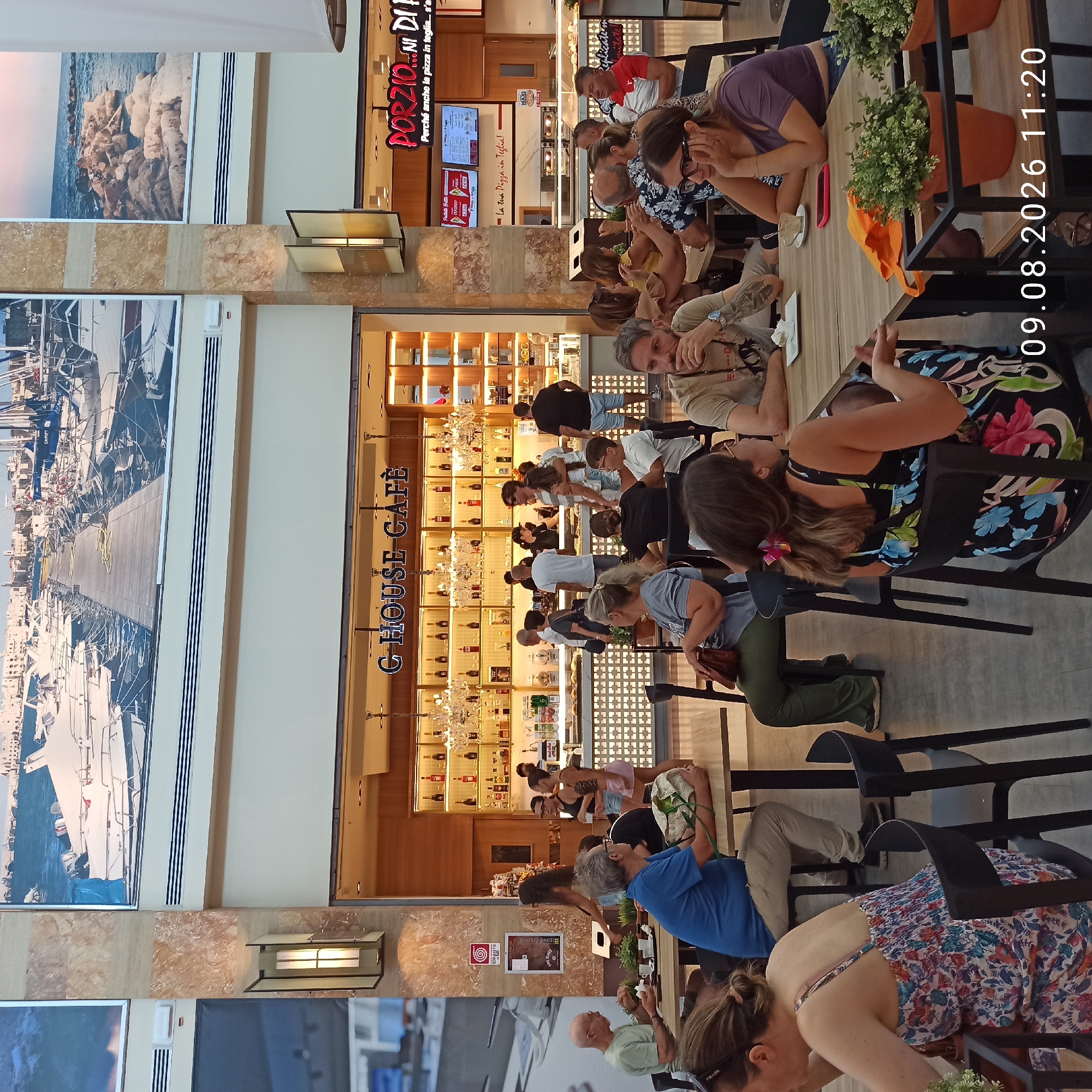 In Molfetta, Italy, within an indoor commercial setting resembling a food court or shopping mall area, several individuals are captured in a moment of leisure. The scene is dominated by the warm, earthy tones of the architecture, with marble-like pillars and a large photographic mural depicting a harbor filled with sailboats adorning the upper walls. Below the mural, the signage for "G HOUSE CAFÈ" is prominently displayed.
In the foreground, patrons are seated at various tables. A woman in a colorful floral dress is seated on the right, facing away from the camera, her hands gesturing as if in conversation. To her left, a man in a grey t-shirt with Japanese characters visible on the sleeve is seated, resting his chin on his hand. Next to him, a woman with glasses is looking downwards, her hand supporting her head. To the left of the woman in the floral dress, a man in a blue shirt and beige pants is seated at a table, his right arm resting on the table. In the middle ground, a group of people are standing and interacting around a cafe counter, which is stocked with bottles and other bar supplies. Behind the counter, staff members are visible. To the right of the cafe, a sign for "PORZIO...NI DI" is visible, suggesting a food establishment. The overall ambiance suggests a casual, public gathering space with a focus on food and drink. The time stamp "09.08.2026 11:20" is superimposed on the lower right portion of the image.