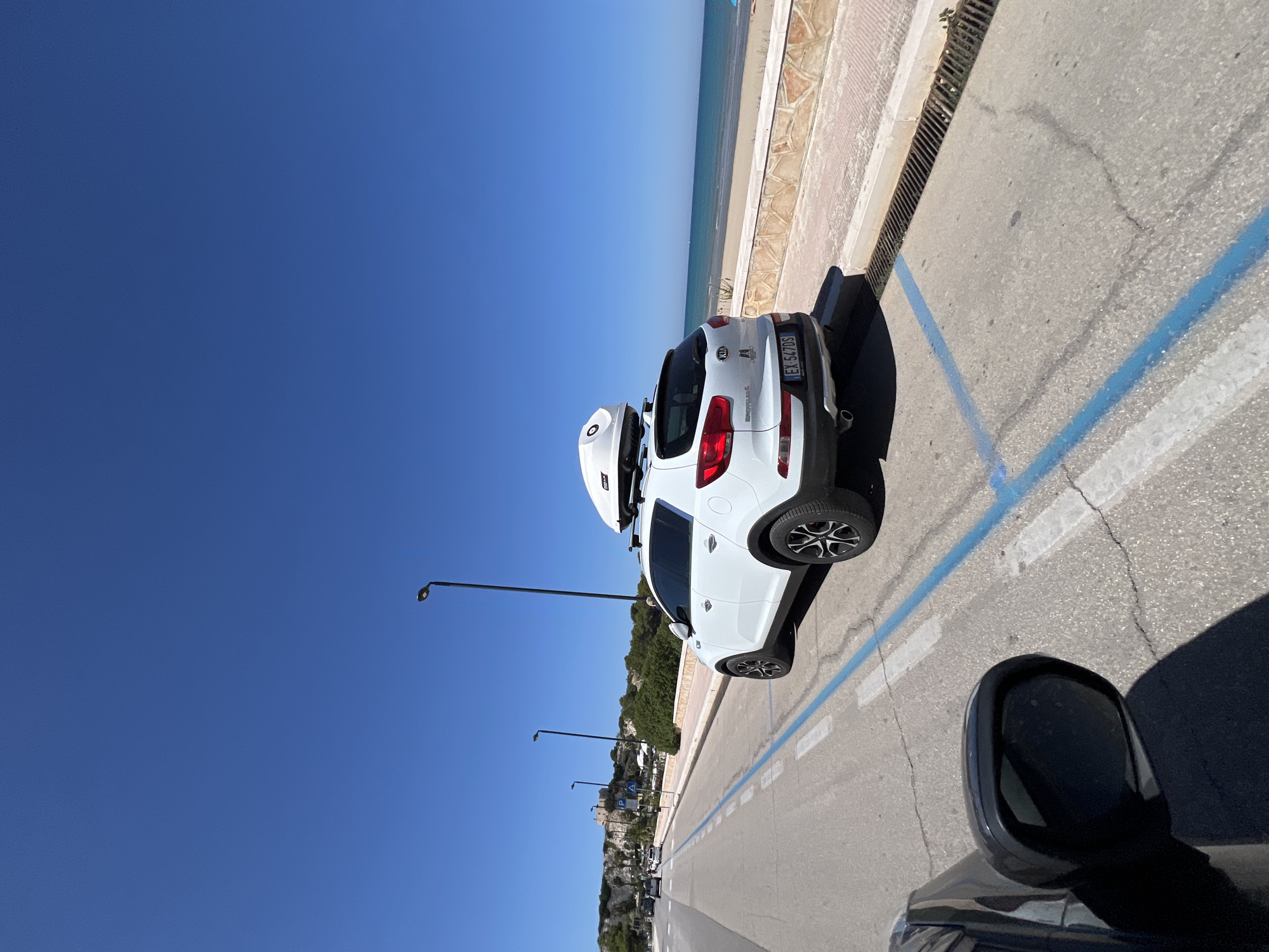 A white Kia Sportage SUV is parked on a street near the coast. The car has a white cargo box on its roof. The scene is a sunny day with a clear blue sky. The street is paved with asphalt and marked with blue and white parking lines. A concrete curb with a metal grate runs alongside the street. Beyond the curb is a tiled walkway and then a sandy beach meeting the ocean. In the distance, there are buildings and lush green trees on a hillside. The image appears to be taken from another vehicle, as a side mirror is visible in the foreground. A license plate on the back of the Kia reads "EE 547DS". The overall impression is a picturesque coastal scene on a bright, warm day, likely in Vieste, Italy, given the location context.