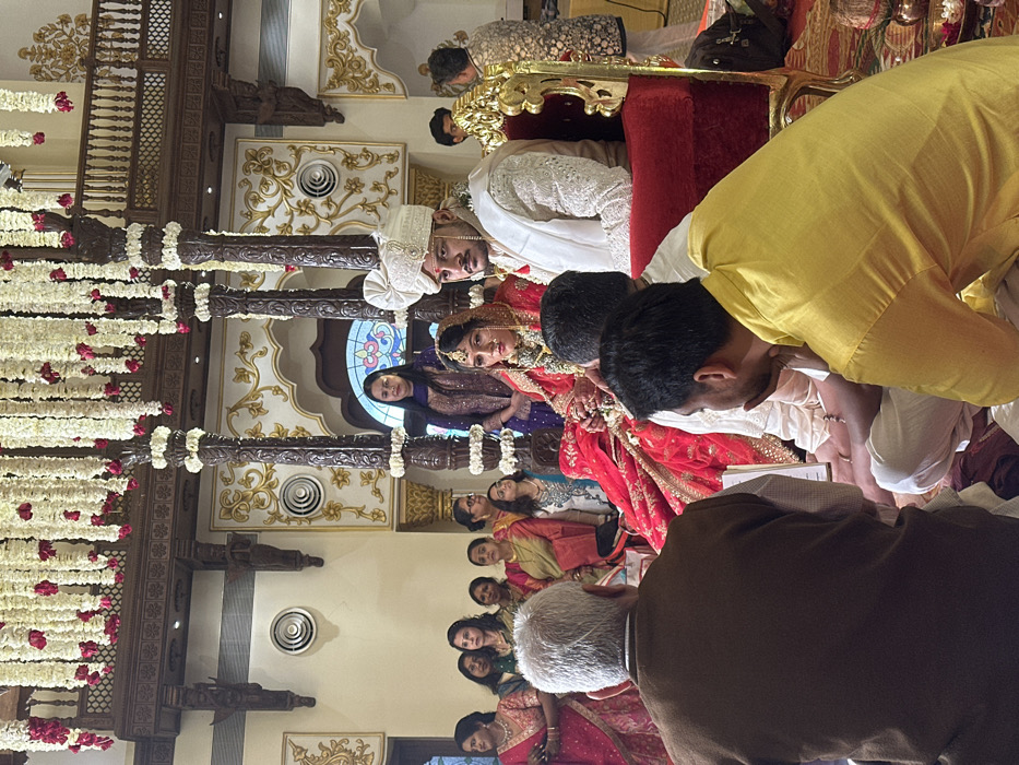 An indoor Hindu wedding ceremony is depicted in a grand hall with traditional Indian architecture, located in Vadodara, India, likely within a palace or heritage venue. The scene is dominated by a central mandap structure featuring four ornate, dark wooden pillars extensively draped with strings of white and red flowers. Hanging floral garlands also descend from the high ceiling, which exhibits intricate wooden beams and carvings.
At the center, the groom is seated on a red velvet chair, wearing a cream-colored sherwani with embroidery, a matching turban, and a pearl necklace. He has a mustache and a focused expression. To his left, the bride sits on a similar chair, adorned in a red lehenga-choli with gold embroidery, elaborate jewelry including a mang tikka and nose ring, and a red veil partially covering her head. Her hands are clasped as if participating in a ritual.
In the immediate foreground, two men are seated on a red carpet with their backs to the camera, facing the couple. The man on the left has grey hair and wears a brown waistcoat over a white shirt. The man on the right wears a yellow kurta and is seen with his right hand near his chin, attentive.
Behind the bride and groom, a woman in a dark purple embroidered outfit stands observing. To the far left, a group of approximately eight women in various traditional Indian attire (sarees, salwar kameez) stand and observe the ceremony. Additional guests are indistinctly visible in the background.
The walls are light-colored with intricate gold and white plasterwork, archways, and traditional motifs. Circular ventilation grilles are visible on the wall. A decorative wooden balcony with intricate railing is present in the upper background, above a large arched window with a stained-glass design. Small metallic ritual objects are visible on the floor in front of the seated men. The lighting is bright, illuminating the festive and formal environment.