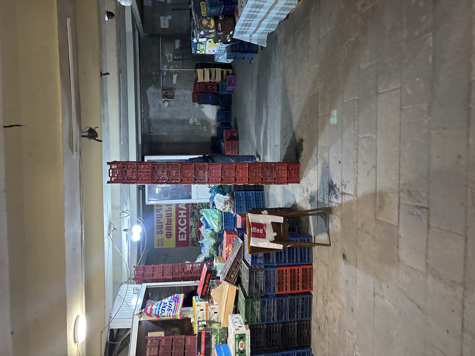 The image captures a scene within a Yerevan, Armenia market or storage area. The location appears to be an indoor market or large indoor stall, identified by the stacked plastic crates and produce. A person is lying between two stacks of red crates. The scene is poorly lit, revealing elements like a chair with a "coffee" sign and various fruits and vegetables stacked in boxes and crates. Several crates of different colors - red, blue, black, and green are also stacked, indicating a variety of goods being stored or sold. A sign, reading "EXCHA", and in Armenian, is also partially visible above the produce stacks. Overhead, there are fluorescent light fixtures providing minimal illumination. The floor is concrete.