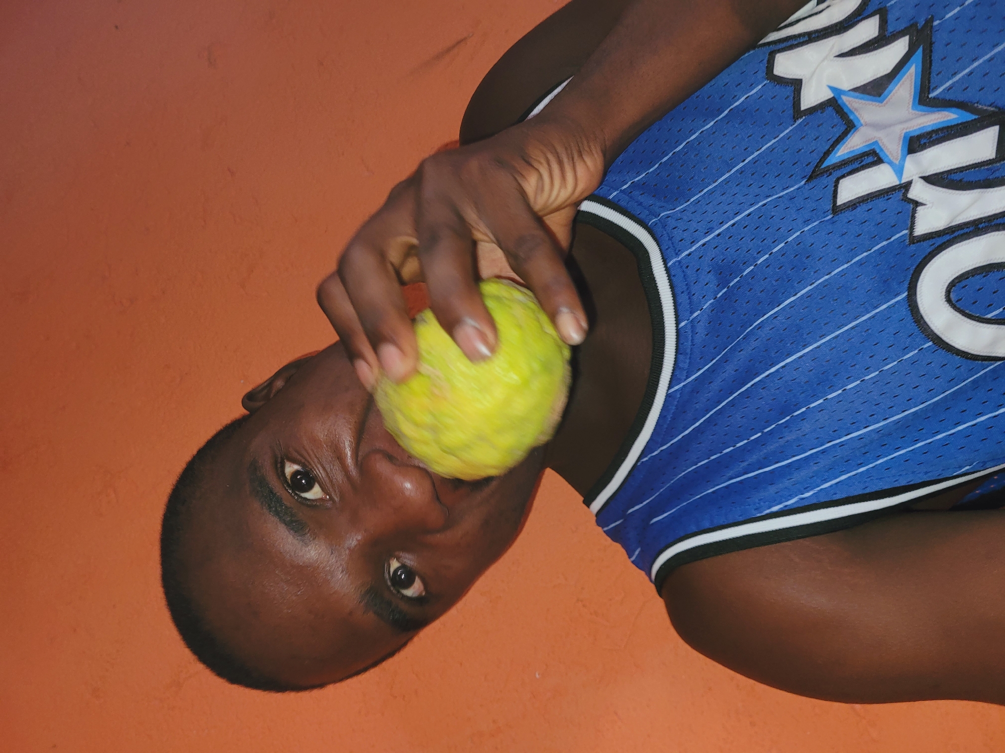 The media file shows a close-up, rotated image of a young Black person, likely male, holding a round, textured yellow fruit, possibly a custard apple or soursop, up to their mouth. The person is wearing a blue basketball jersey with white pinstripes and a white and blue star logo on the chest. The jersey has white and black trim around the neck and armholes. The background is a textured orange wall. The person's face is visible, with their eyes open and looking towards the camera. Their expression is neutral. The image appears to be taken indoors, with no clear indicators of the time of day or weather. The location is identified as Bukuru, Nigeria. There is no visible text in the image. The lighting is somewhat harsh, casting shadows on the person's face and arm. The focus is on the person's face and the fruit they are holding.