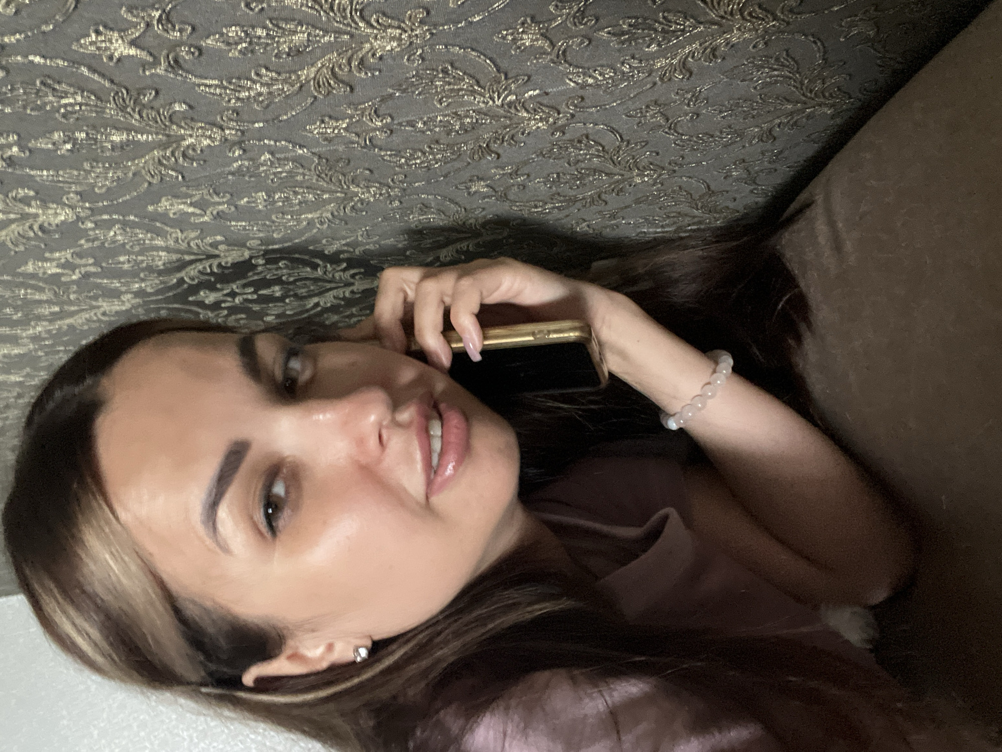 A woman with long, dark brown hair is lying down, speaking on a gold-colored mobile phone. She is wearing a light pink shirt and a bracelet with clear beads on her left wrist. Her right hand is holding the phone to her ear. The background is a textured wallpaper with an ornate, damask pattern in shades of gray and gold. The lighting is dim, with a warm glow emanating from the phone and casting shadows on the woman's face and arm. The setting appears to be indoors, possibly a bedroom or living room.