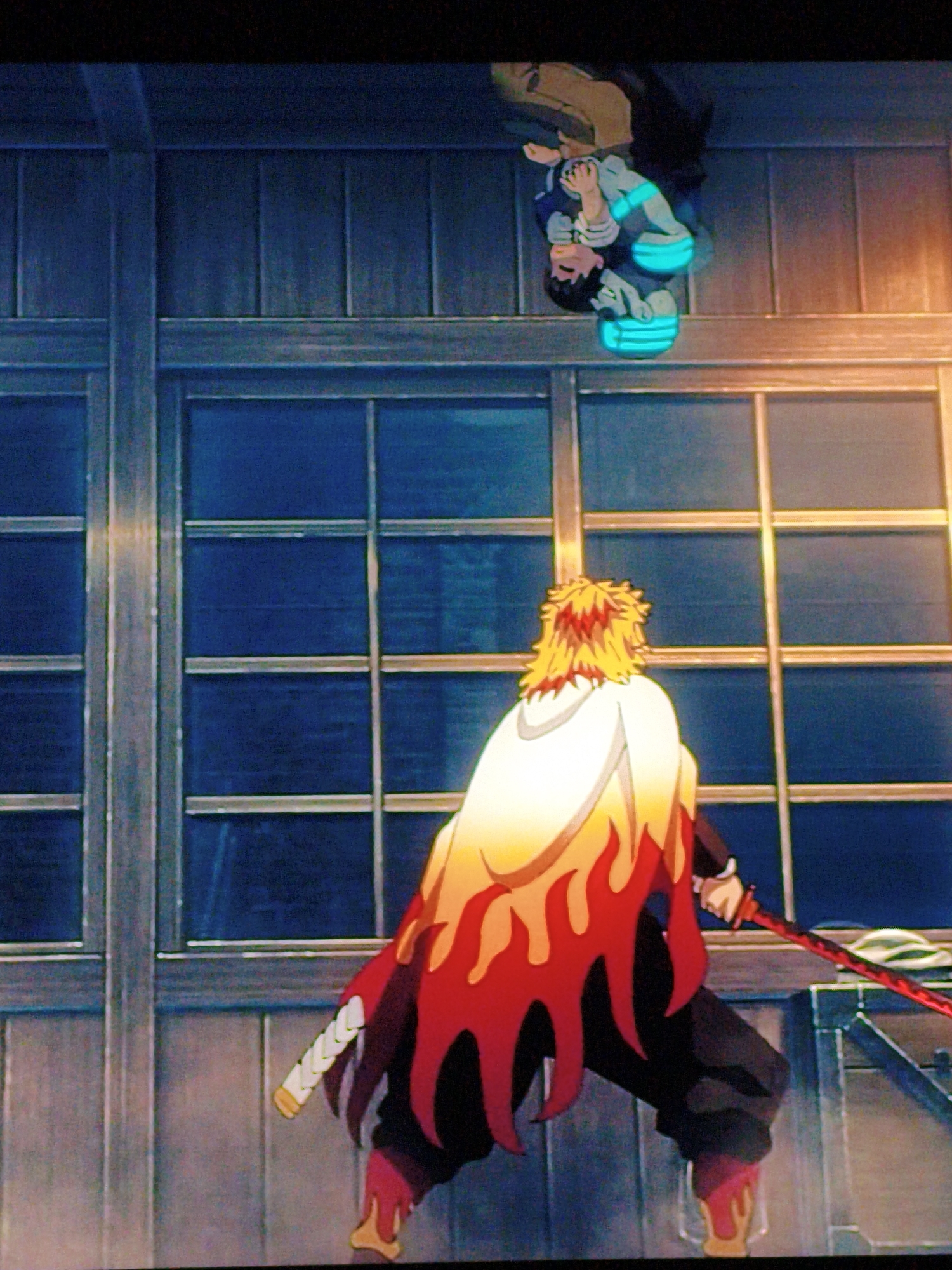 The image depicts an animated scene with two characters. In the foreground, a character with spiky blonde hair and a white cloak adorned with flame patterns is facing away from the viewer, holding a red-bladed sword. They are wearing dark clothing. In the background, above and to the left, another character is depicted upside down, wearing a blue and white outfit with glowing turquoise accents on their legs. This character appears to be suspended in mid-air. The environment seems to be indoors, with large windows featuring a grid pattern and wooden paneling on the walls. The lighting suggests it is either dusk or night. There is no discernible connection to Vladivostok, Russia.