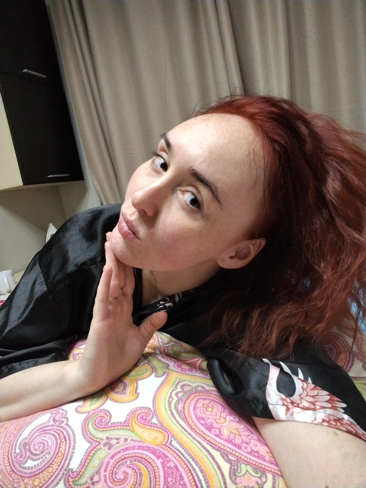 An adult female with reddish-brown, wavy hair and fair complexion is depicted in a close-up, angled self-portrait. She is positioned on a surface covered with a pink, yellow, and green paisley-patterned fabric, likely a bedspread or cushion. Her right hand, with a plain ring on the ring finger, is placed under her chin. She wears a black, satin-like garment, possibly a robe, with a white and pink/brown patterned section visible on the lower right sleeve. Her dark eyes are directed towards the camera. The background on the left reveals a dark brown cabinet with light-colored handles, and partially visible white objects resembling a mug and an iron. To the right, a beige curtain hangs with vertical folds. The scene is indoors, indicative of a residential environment. The image contains no discernible visual elements that would identify a specific location within Izhevsk, Russia.