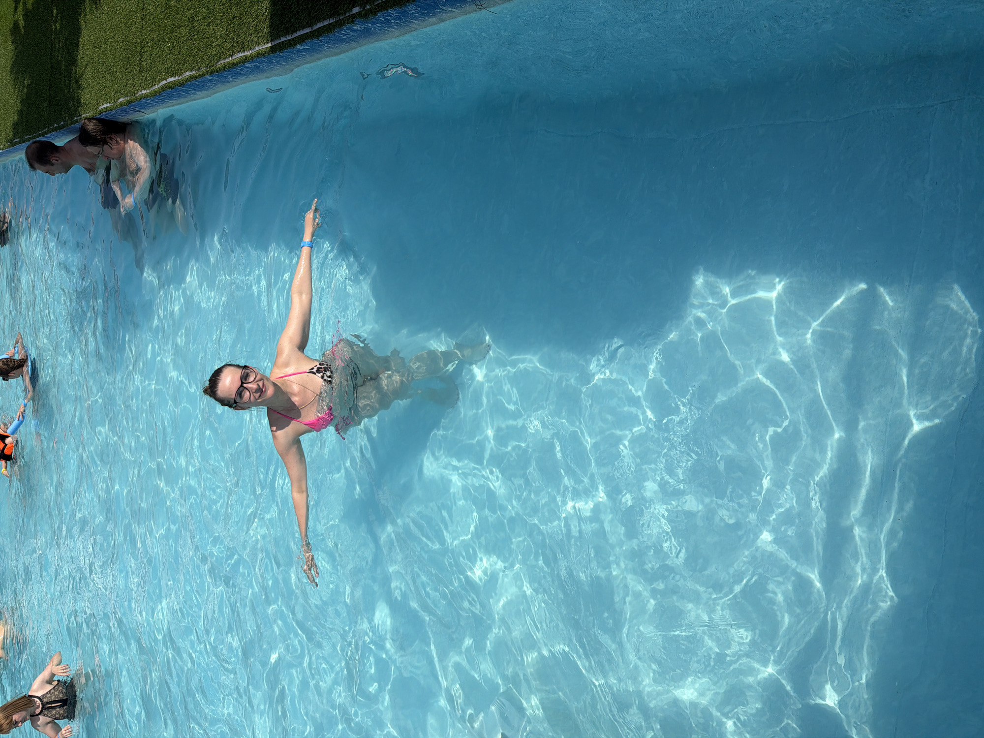 A woman in a swimsuit is swimming in a pool. She is wearing glasses and a bracelet. Several other people are in the pool. The pool has blue water and is located next to a patch of artificial turf. The location of the pool is not discernible from Moscow, Russia.