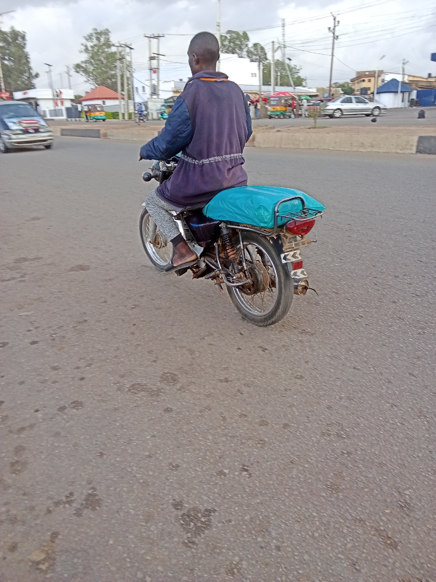 An individual is riding a motorcycle on a paved road in Gura Top, Nigeria. The person is wearing a purple jacket and light gray pants, with sandals on their feet. The motorcycle has a turquoise cover on the back and a visible exhaust pipe. In the background, there are other vehicles, including a silver car and a green and yellow motorized tricycle, as well as buildings, trees, and utility poles. The sky is overcast.