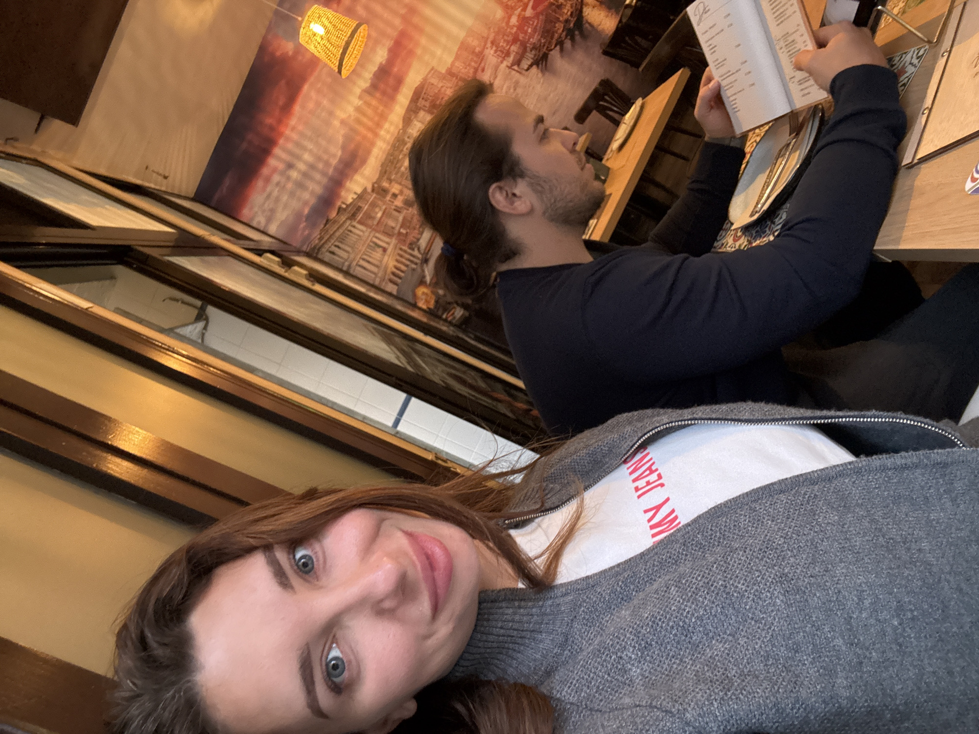 A selfie taken at a restaurant in València, Spain. A woman with auburn hair and blue eyes smiles at the camera. She wears a grey sweater over a white t-shirt with red lettering. Behind her, a man with long brown hair is seated at a table, reading a menu. The table is set with plates and cutlery. Behind the man is a wall featuring an image suggestive of a cityscape. The room has a warm, yellow light, and windows can be seen in the background.