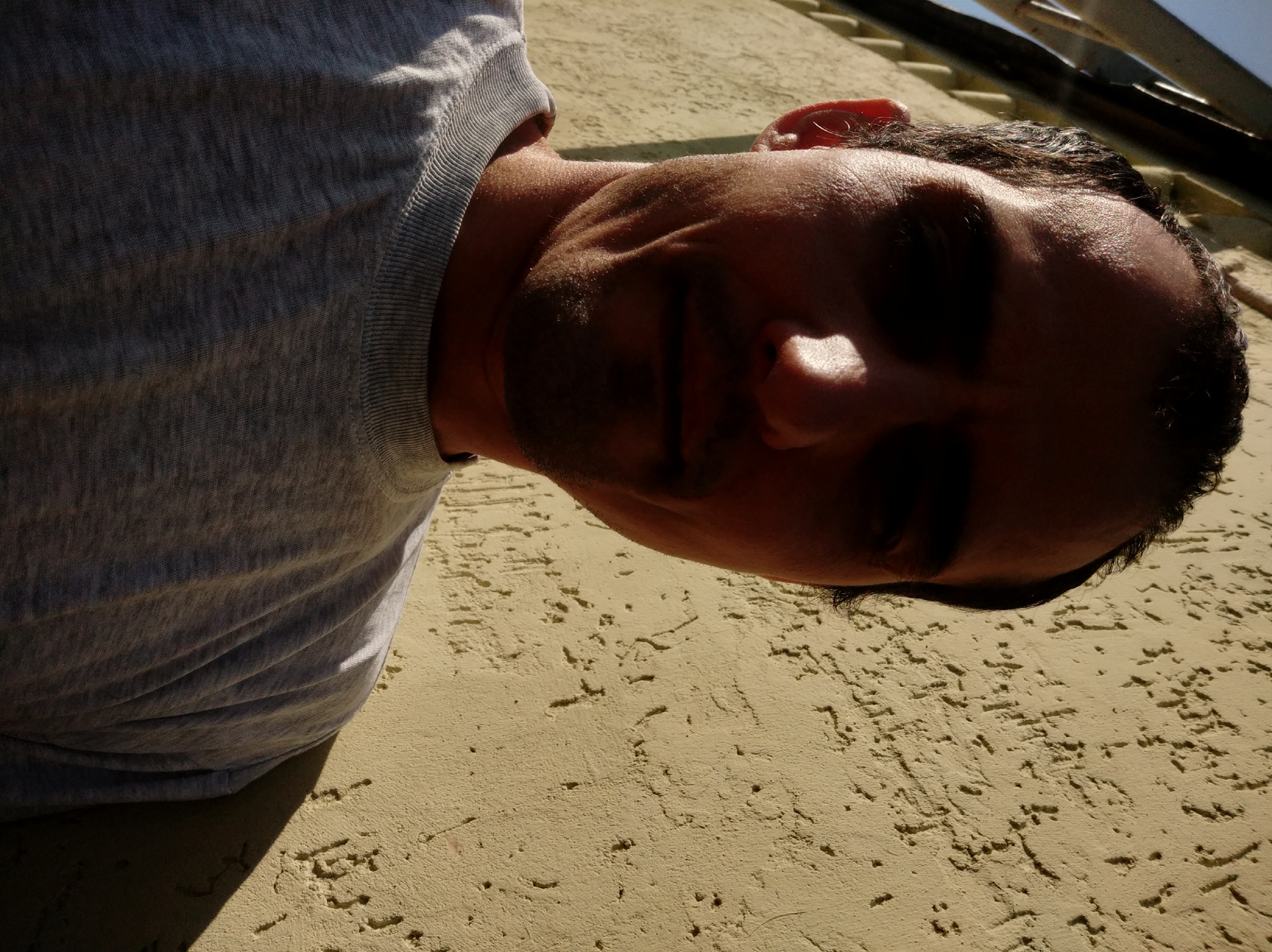 A man is photographed from the chest up, leaning against a textured, yellow wall. The lighting suggests it is daytime, with harsh shadows casting across his face and the wall, indicating a strong light source, likely the sun, to the man's left. The man is wearing a grey, crew-neck t-shirt. His face is partially obscured by shadow, but his forehead, nose, and mouth are visible. His hair is dark and appears short. The background is dominated by the textured wall and some metallic structures in the upper left corner of the image. The overall impression is a casual, outdoor setting, possibly in an urban environment.