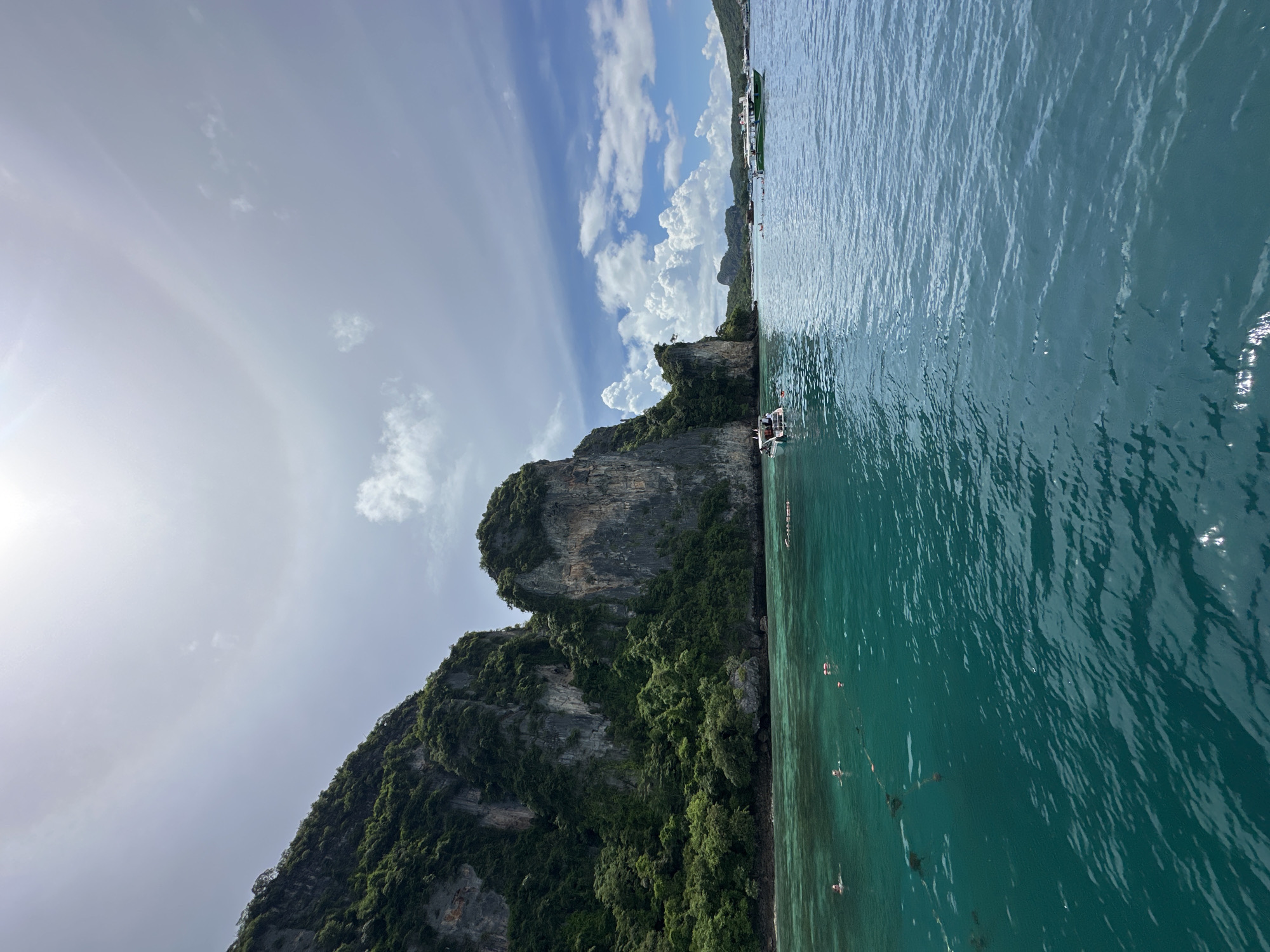 A wide shot captures a vibrant scene in Ao Nang, Thailand, where majestic limestone cliffs draped in lush greenery rise dramatically from the crystal-clear turquoise waters of the Andaman Sea. The sun, partially obscured by a halo-like atmospheric phenomenon, casts a bright glow across the expansive sky, dotted with fluffy white clouds.
Several boats are visible on the water. A small white vessel is anchored near the cliffs, with people swimming and wading in the shallows around it. Further out, a larger green and white boat sails on the sea, and in the distance, more boats and landforms suggest a bustling coastal area. A rope with buoys stretches across the water, marking a boundary. The sea is calm, with gentle ripples reflecting the sunlight. The overall impression is one of a picturesque tropical paradise, ideal for leisure and exploration.