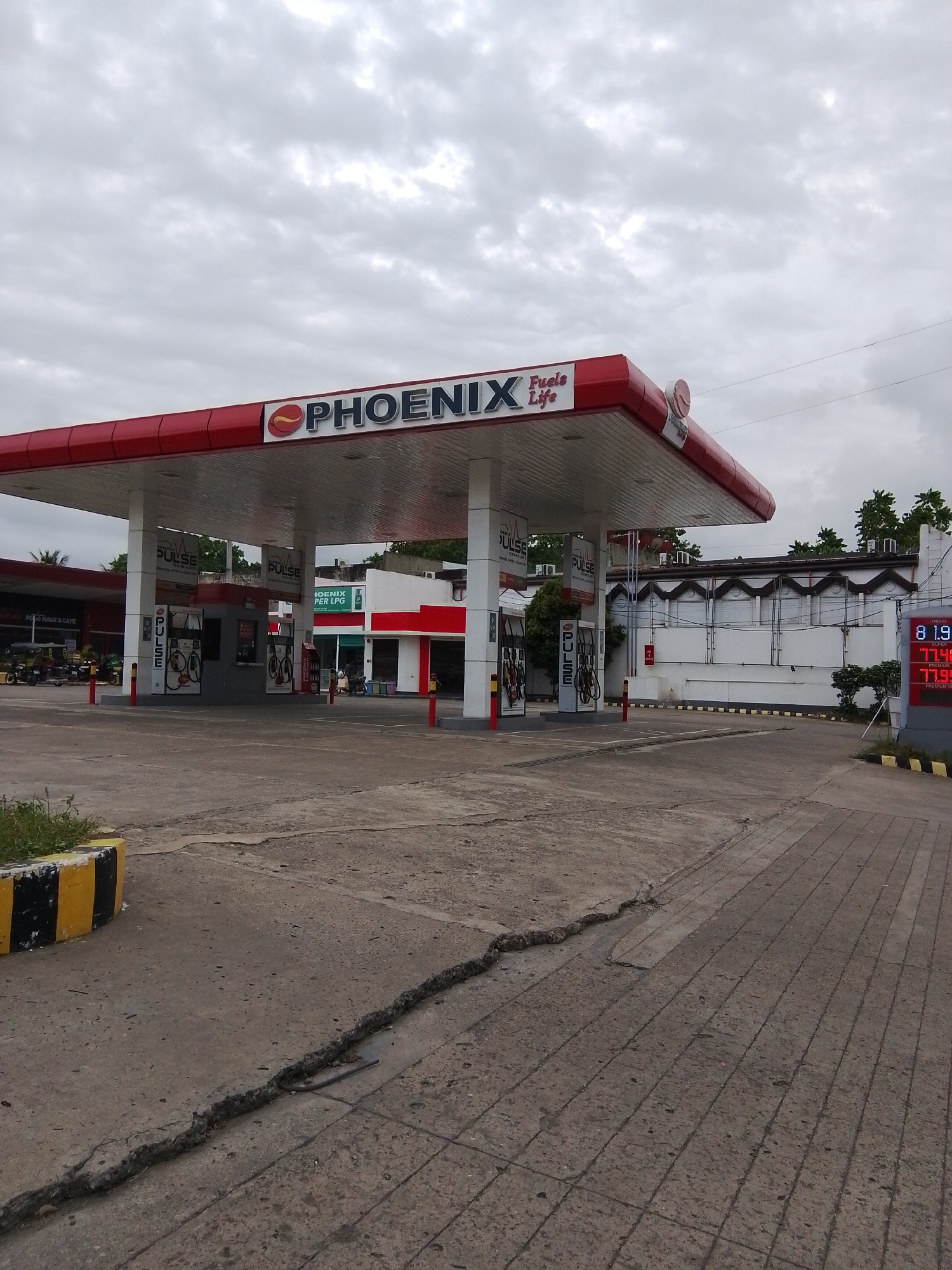The image shows a Phoenix gas station in Davao City, Philippines. The gas station has a red roof and white pillars. There are several gas pumps under the roof. A digital sign displaying fuel prices is visible on the right side of the image. The sky is cloudy. The ground in front of the gas station is paved with concrete, and there is a large crack in the pavement in the foreground. A yellow and black striped barrier is visible on the left side of the image. Buildings are visible in the background, as well as some trees and a few vehicles, including a tricycle.